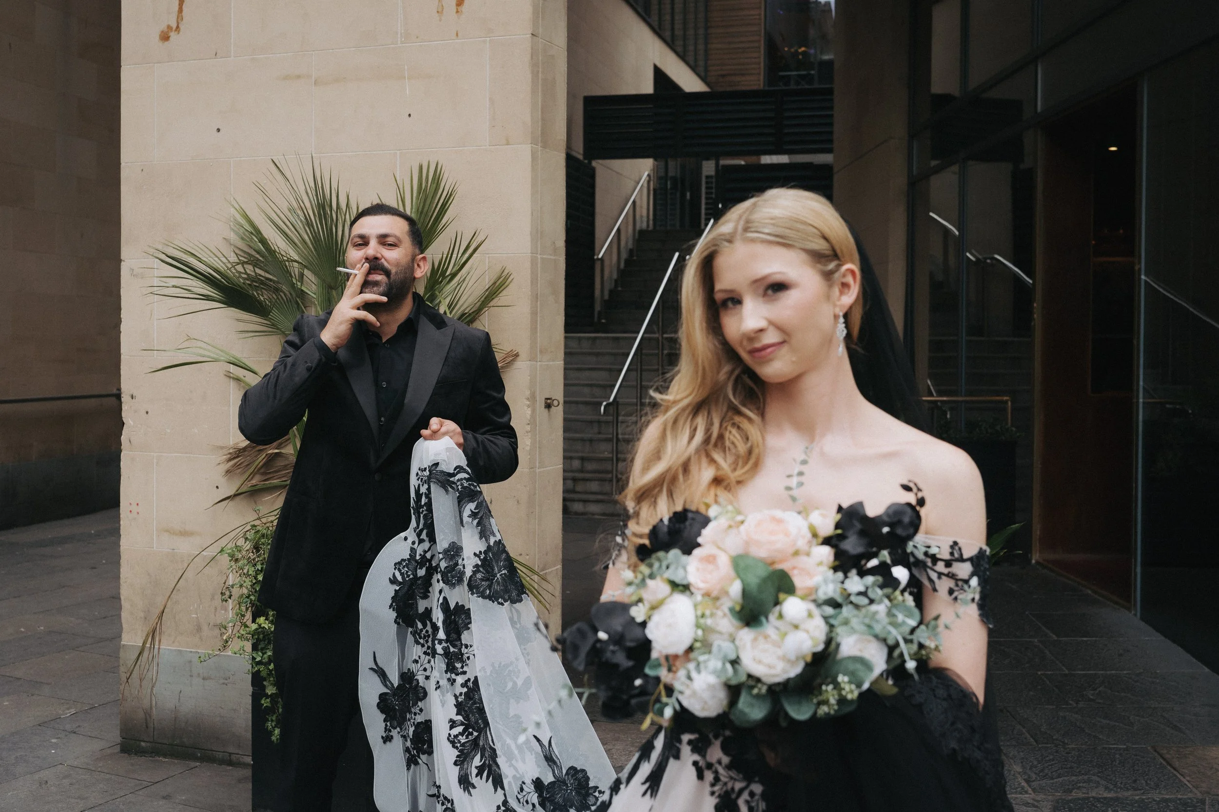 A woman in a black and white dress holding a bouquet of flowers, standing outside a building. A man in a black tuxedo with a black shirt behind her, smoking a cigarette, holding a black and white floral garment. - captured by an Edinburgh wedding pho