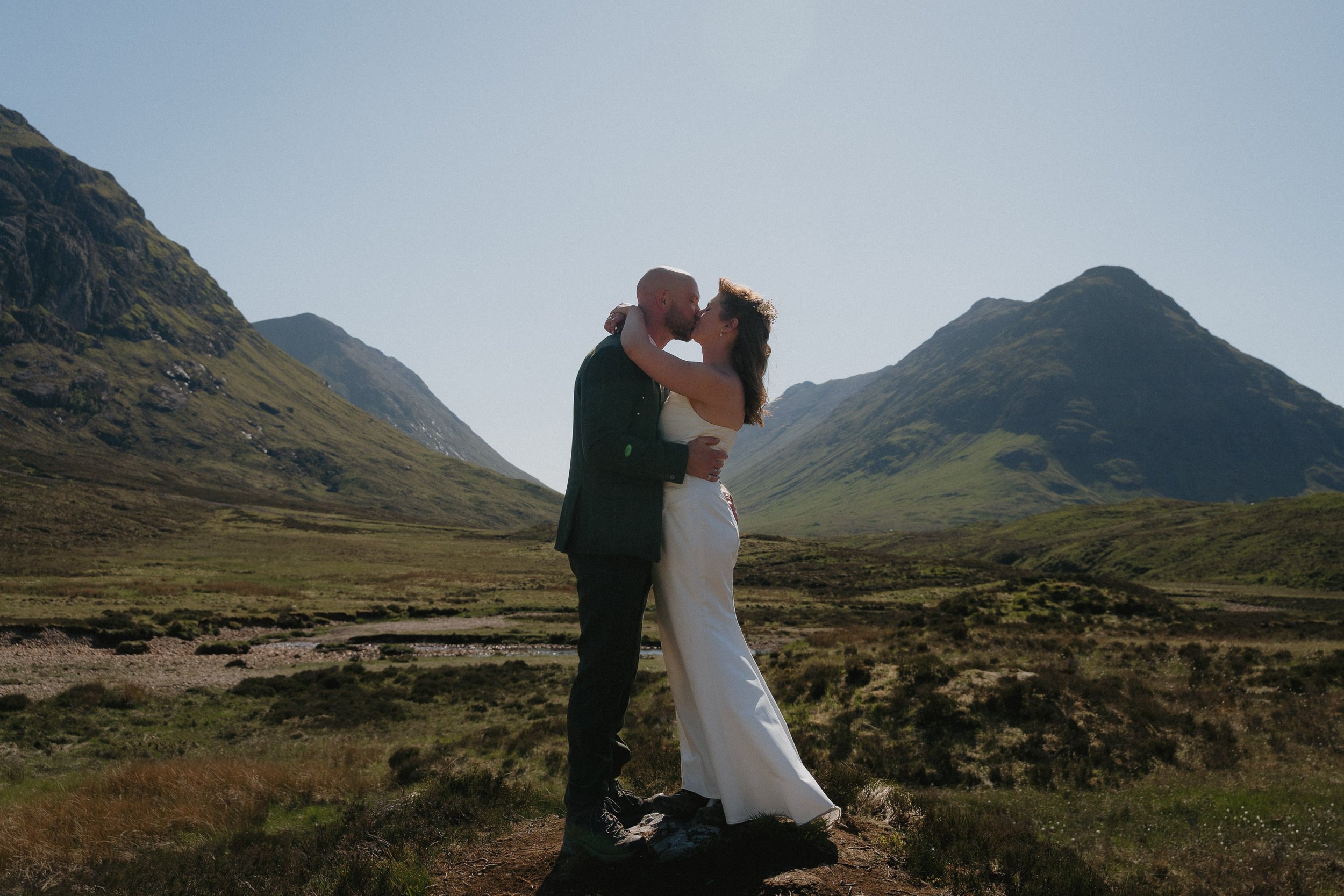 A couple kissing outdoors in a mountainous landscape with green hills and mountains in the background. - captured by an Edinburgh wedding photographer