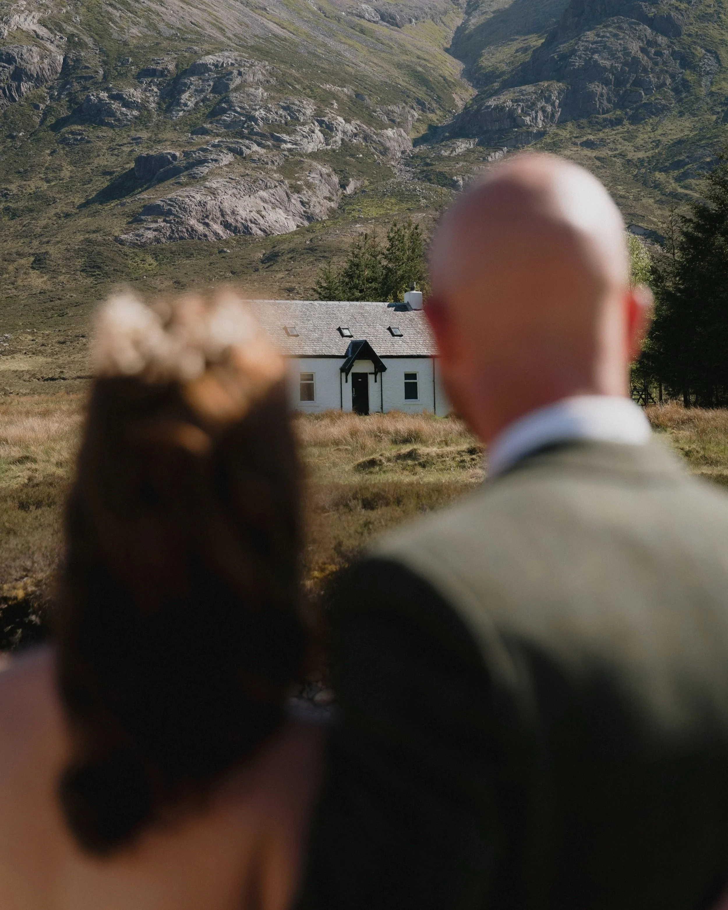 A man and a woman sitting outdoors, facing a house with a mountain backdrop  - captured by an Edinburgh wedding photographer