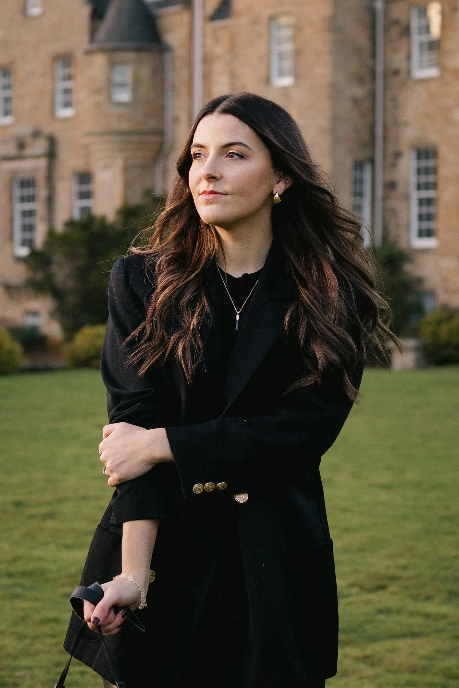 A woman in a black blazer standing outdoors on a grassy area with a large, historic stone building in the background.