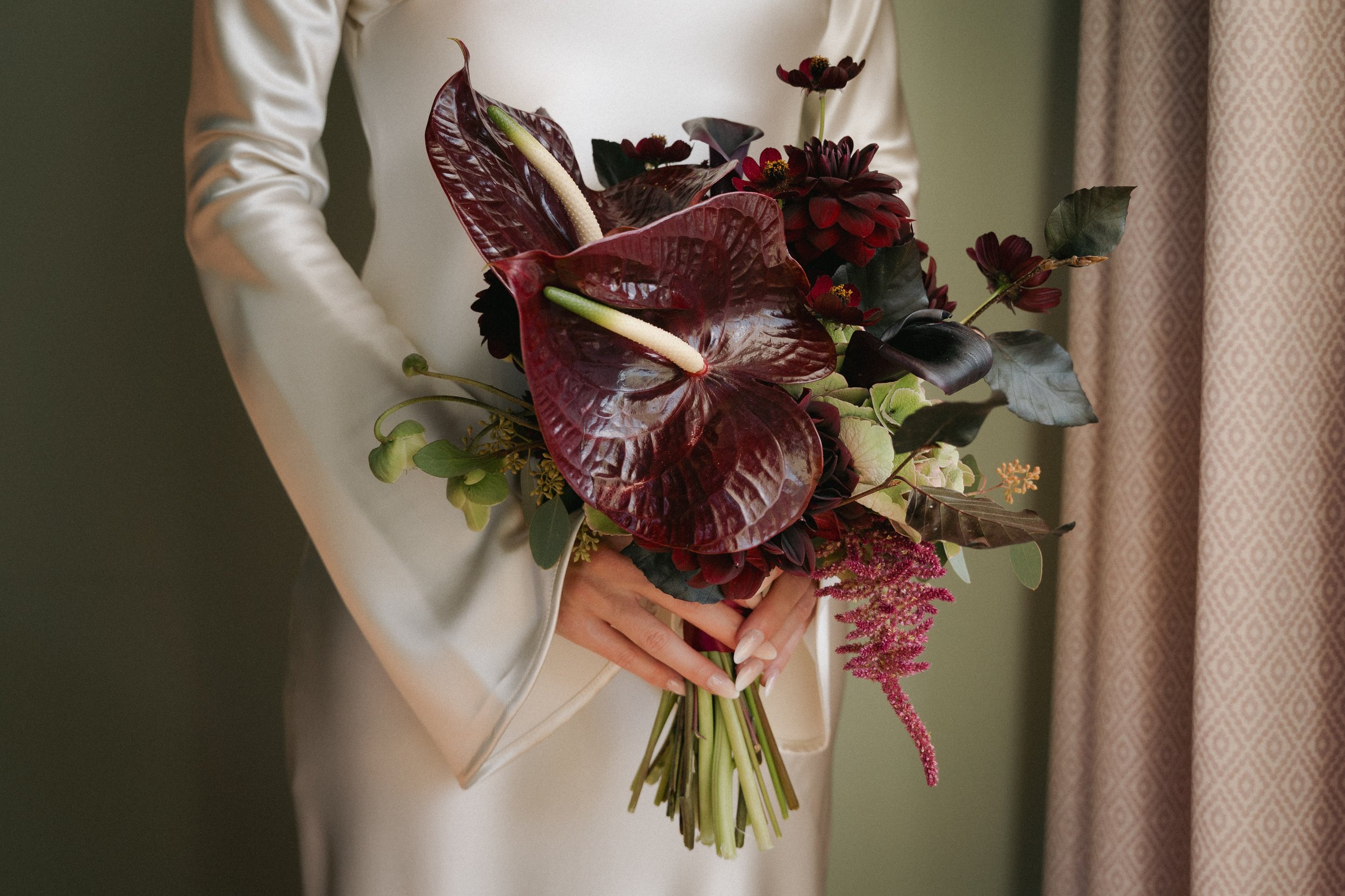 Person in a white satin dress holding a bouquet of dark red and green flowers, including anthuriums, dahlias, and other foliage, against a green and patterned curtain background. - captured by an Edinburgh wedding photographer