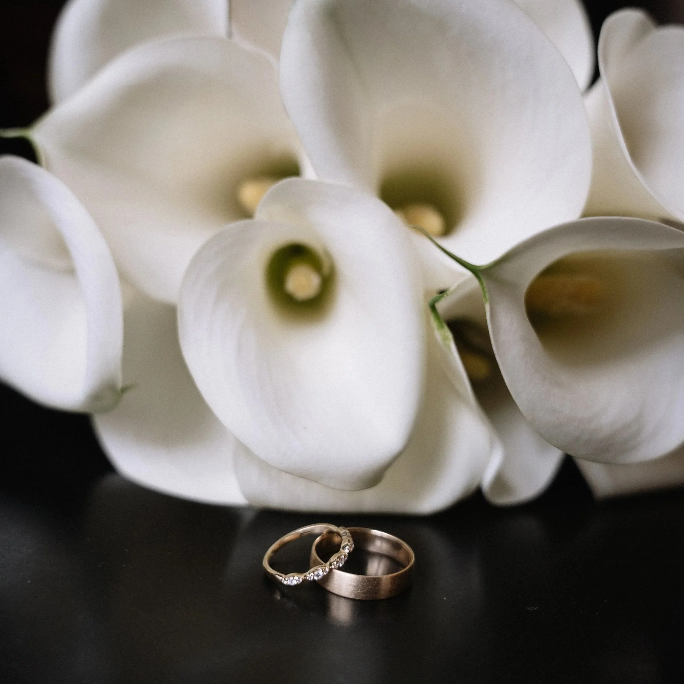 Close-up of white calla lily flowers with two rings, one gold with diamonds, placed on a black surface.