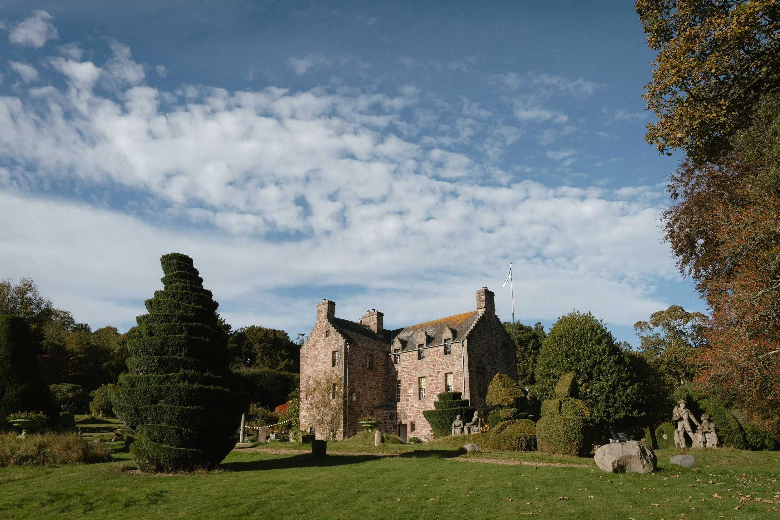 A historic stone building with multiple chimneys surrounded by a manicured garden with shaped trees and sculptures, under a partly cloudy sky. - captured by an Edinburgh wedding photographer