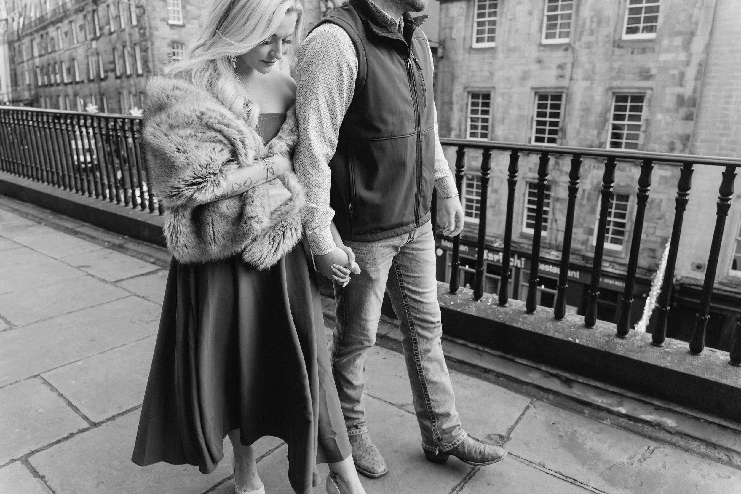 A woman in a gown and fur stole and a man in jeans and a vest are walking hand in hand on a city sidewalk, seen from the waist down - captured by an Edinburgh wedding photographer