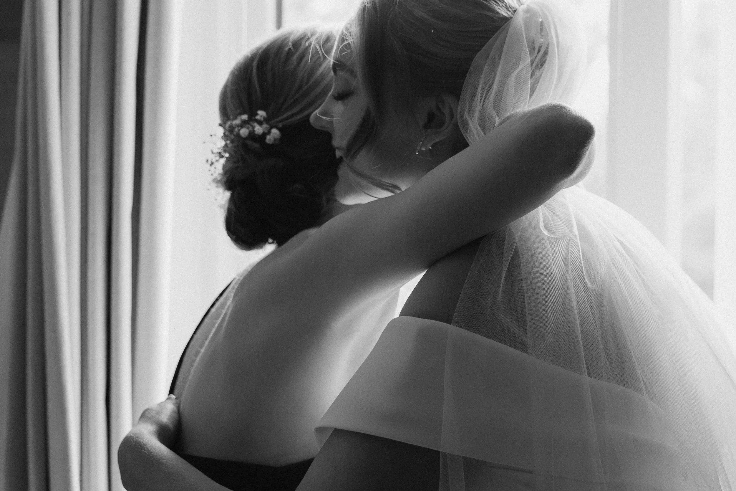 Two women, possibly a bride and her daughter or maid of honor, embrace warmly. The bride wears a white wedding dress and veil, with floral hair accessories, while the other woman has her back to the camera, wearing dark attire. They share a tender mo