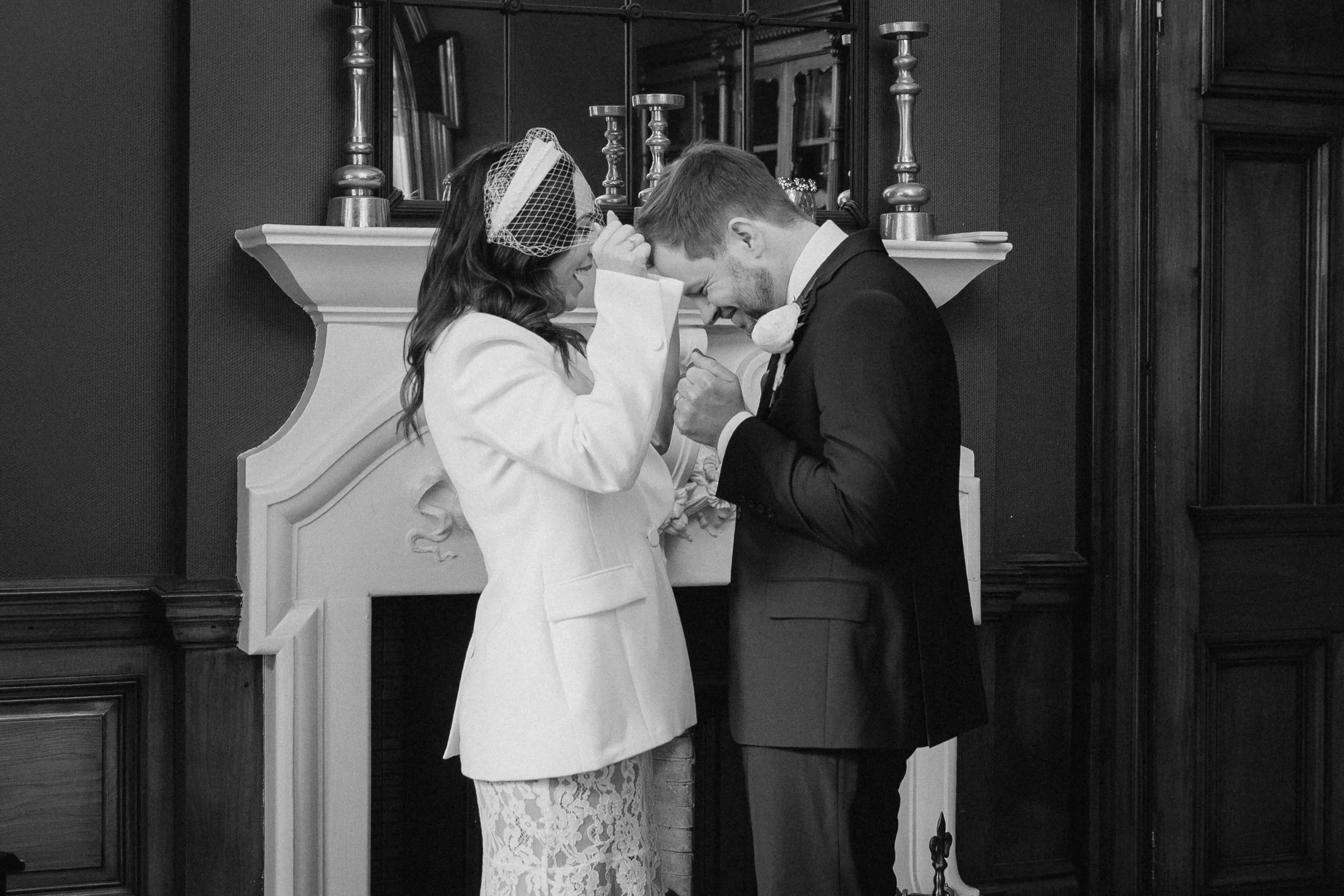 A black-and-white photo of a couple at their wedding, with the woman wearing a white dress with lace details and a veil, and the man in a dark suit and tie, touching foreheads in a tender moment indoors - captured by an Edinburgh wedding photographer