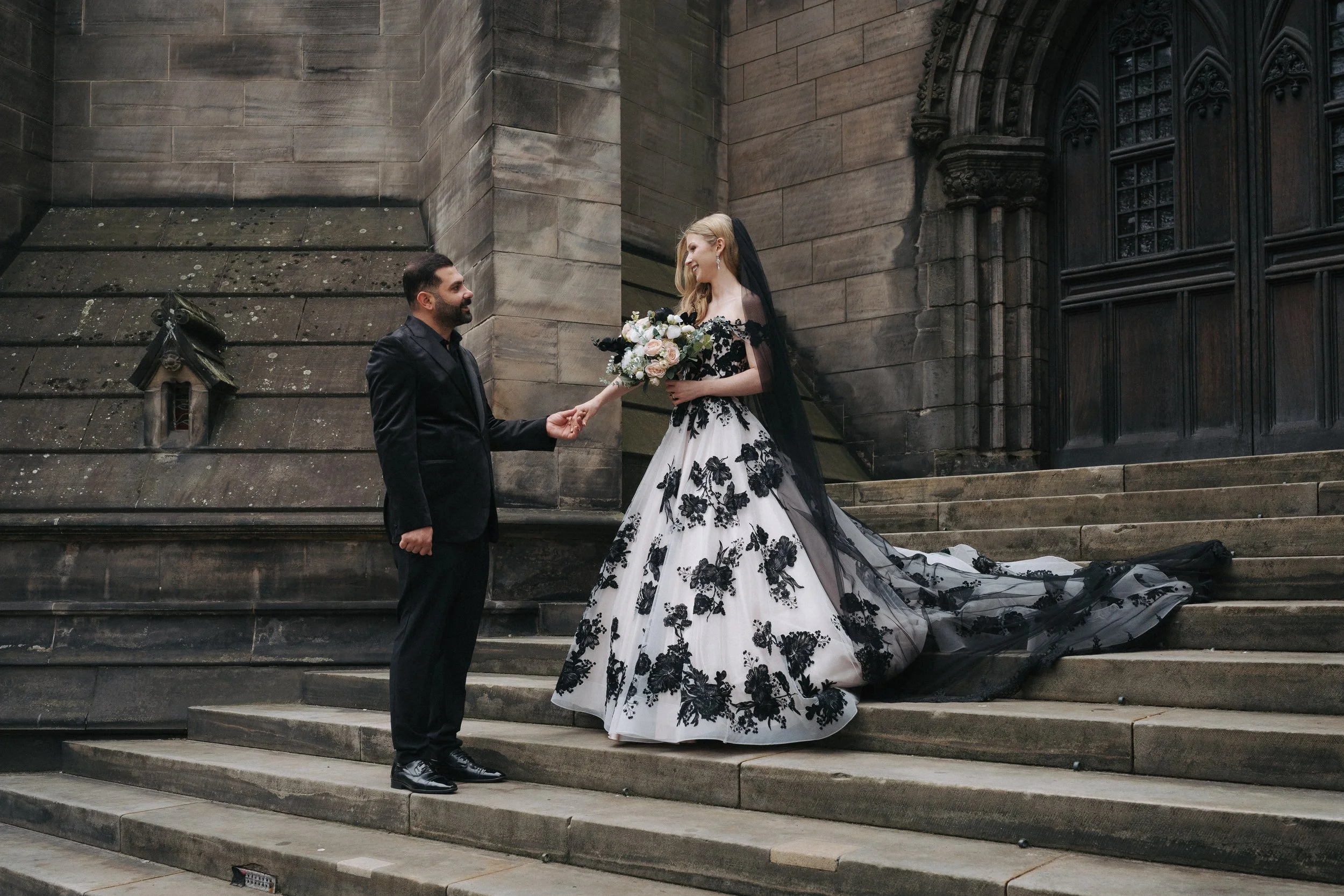 A bride and groom standing on the steps outside a stone church. The bride wears a white wedding gown with black floral lace overlay and a black veil, holding a bouquet of white and pink roses. The groom wears a black tuxedo, holding the bride's hand 