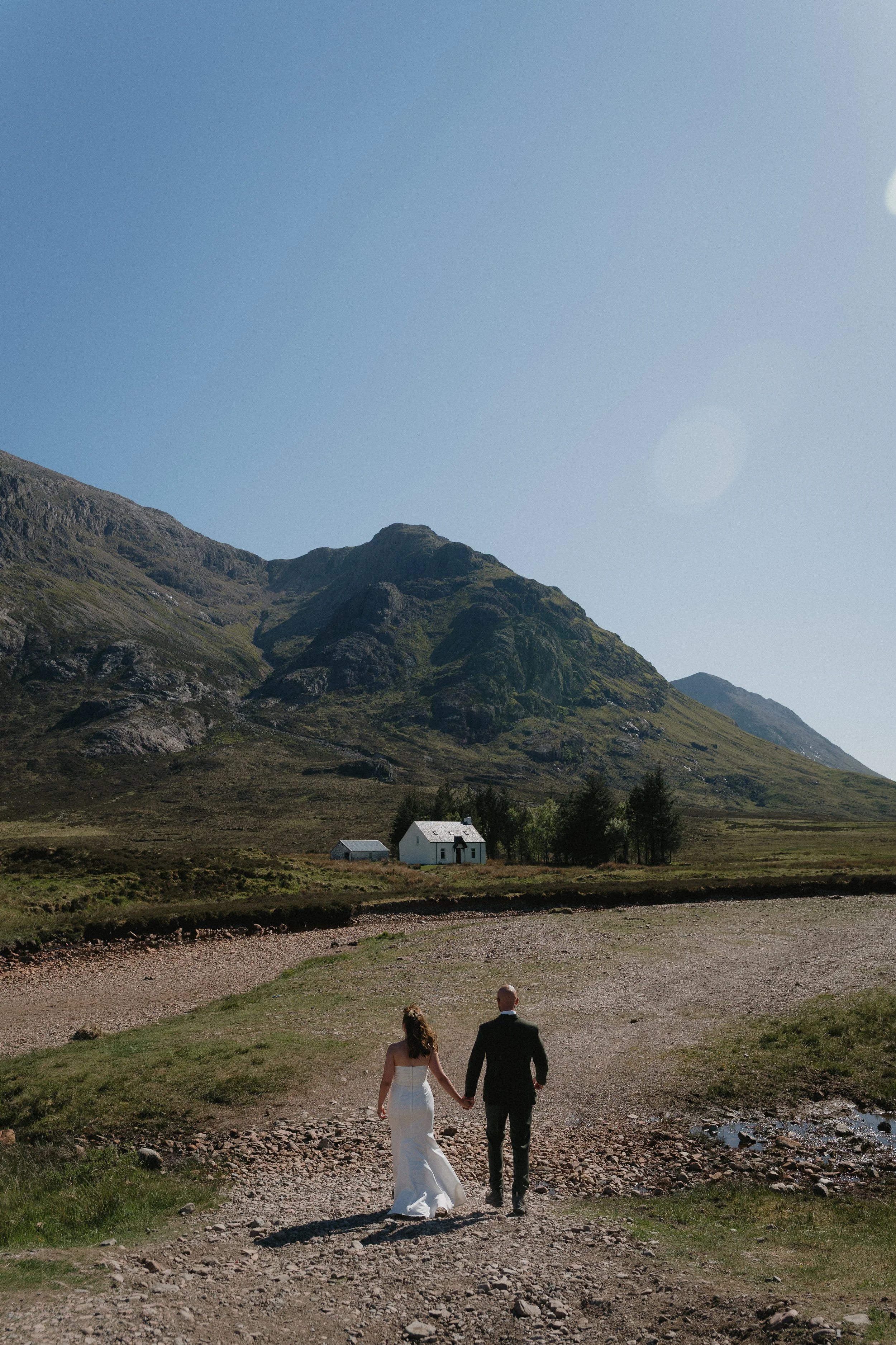 A couple holding hands, walking toward a small white house with a gray roof in a mountainous landscape under a clear blue sky. - captured by an Edinburgh wedding photographer