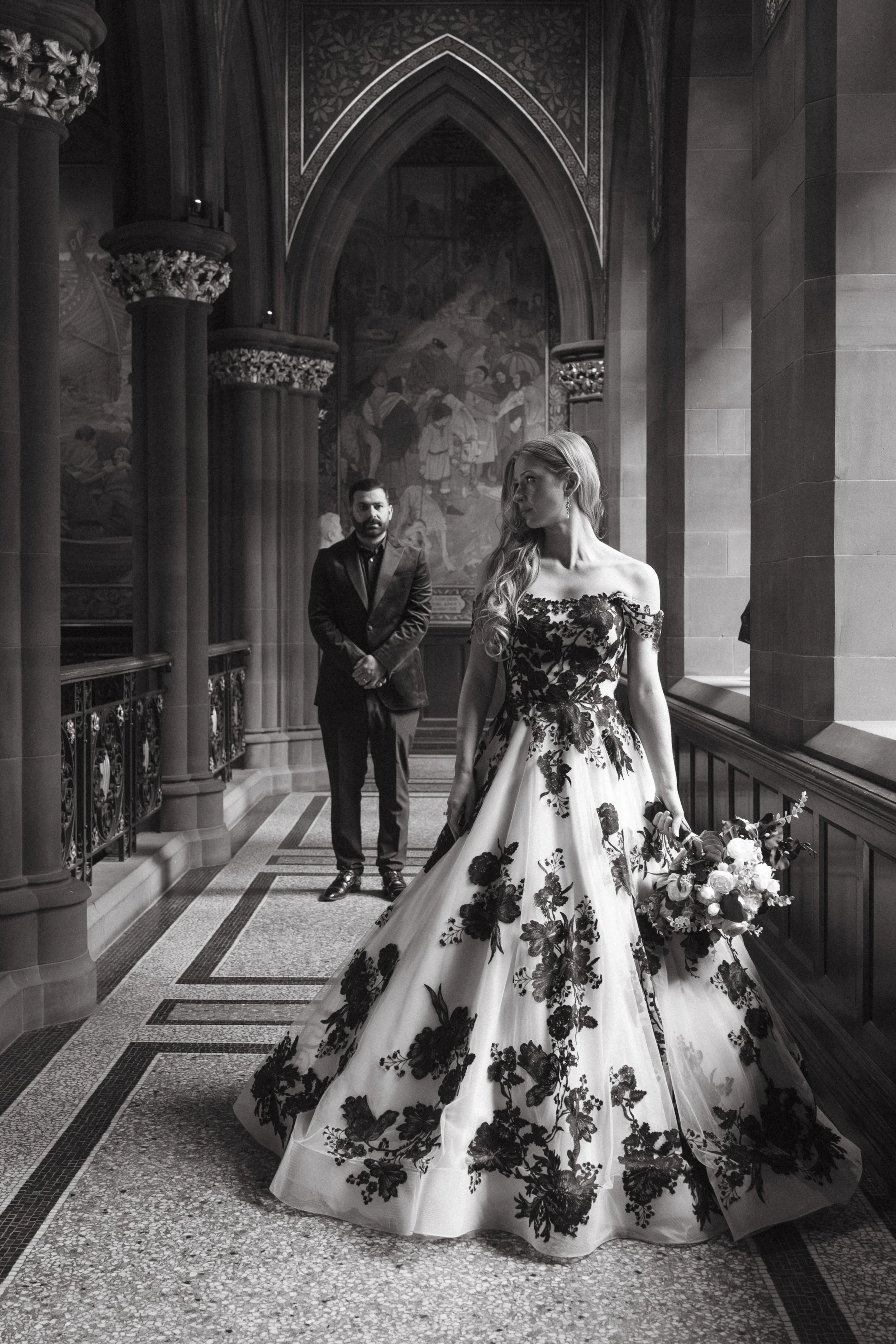 A black-and-white photograph of a bride in an elegant floral wedding gown holding a bouquet, standing in front of a window with stained glass, with a groom in a dark suit in the background in a historic, ornate hallway.