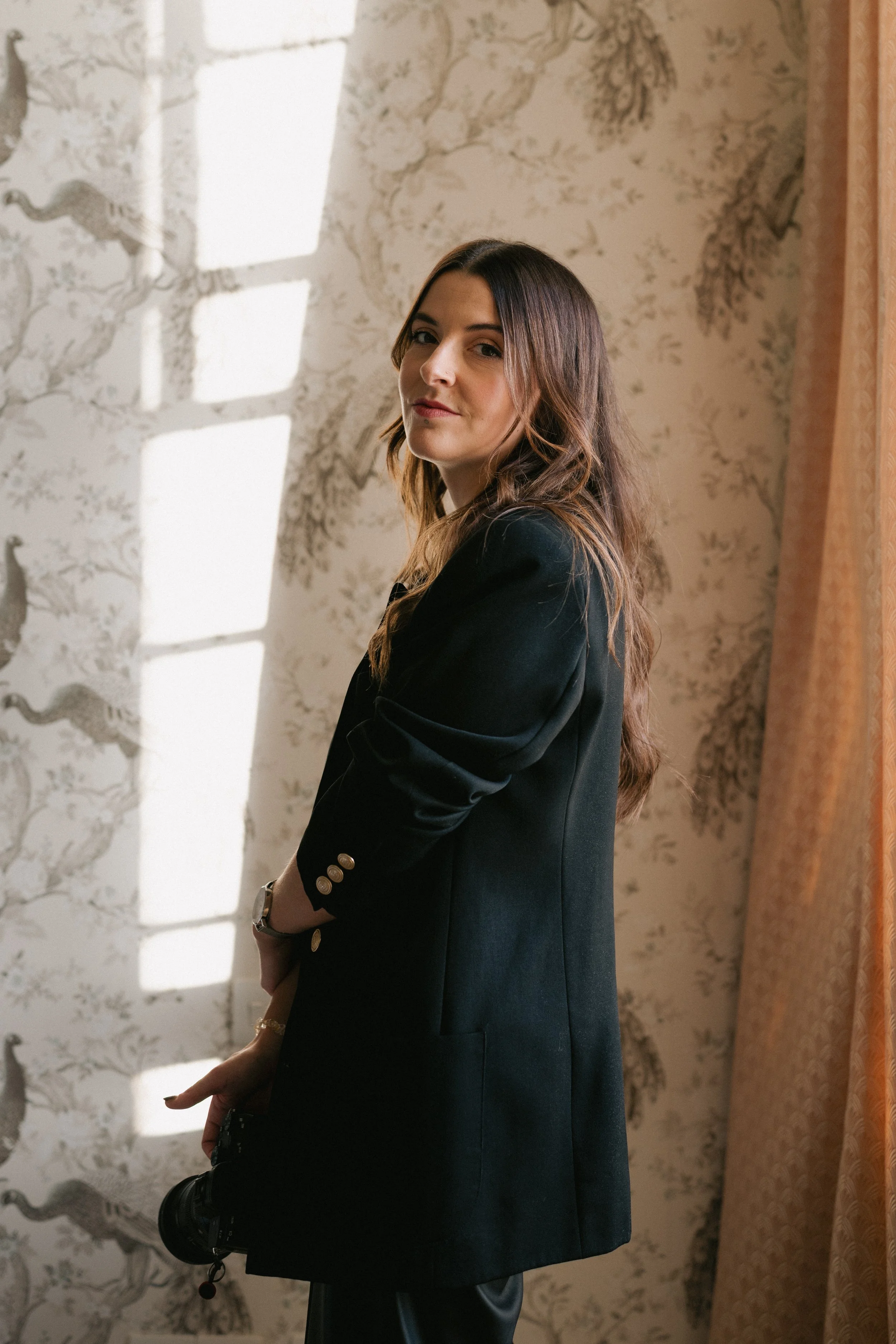 A woman with long brown hair standing indoors, wearing a black blazer with gold buttons. She is holding a camera in her right hand. The background has patterned wallpaper with a nature scene and a window with sunlight streaming in.