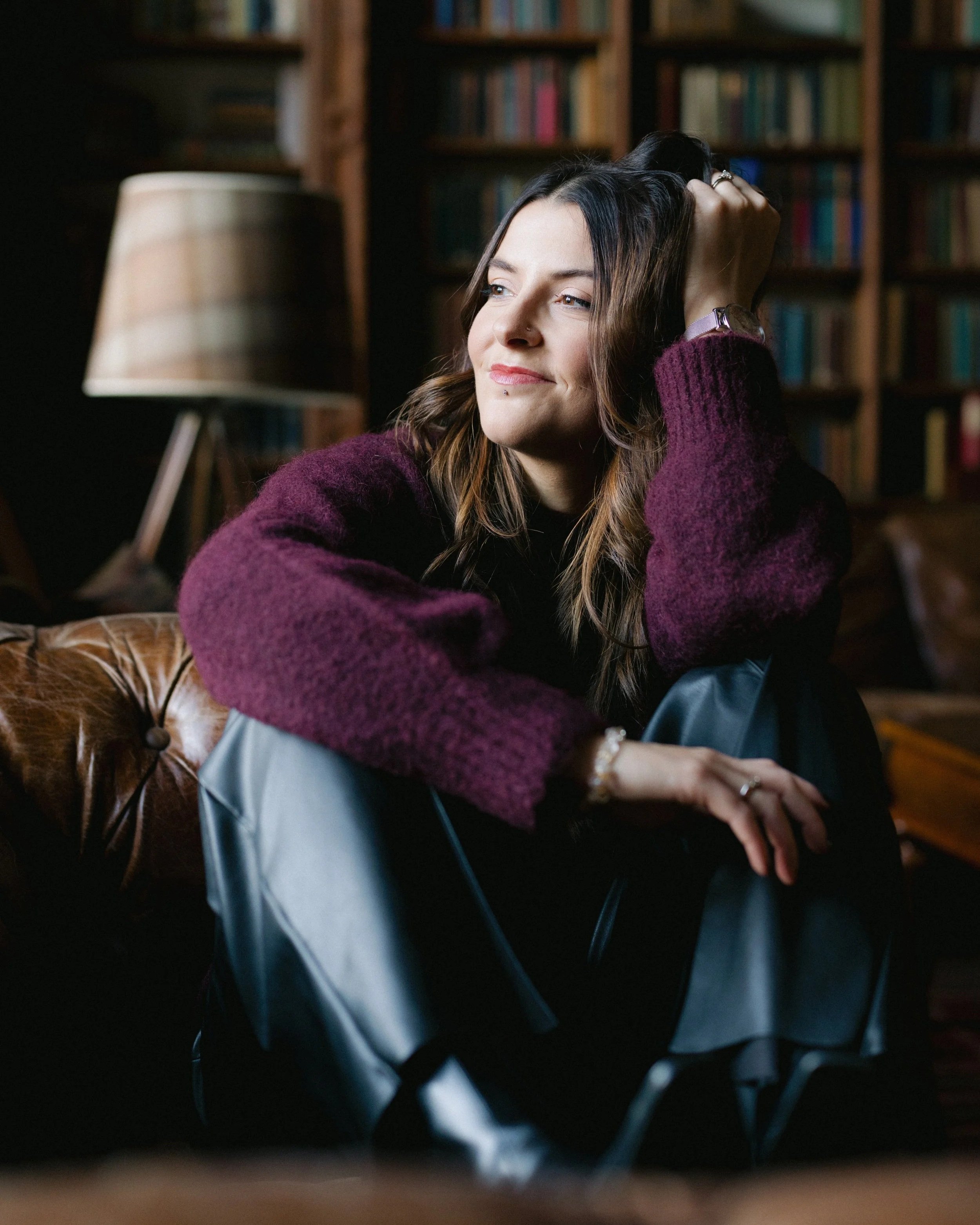 A woman with brown hair and a nose piercing sitting on a leather couch in a cozy room with a bookshelf in the background. She is wearing a purple sweater and black pants, looking thoughtfully to the side.