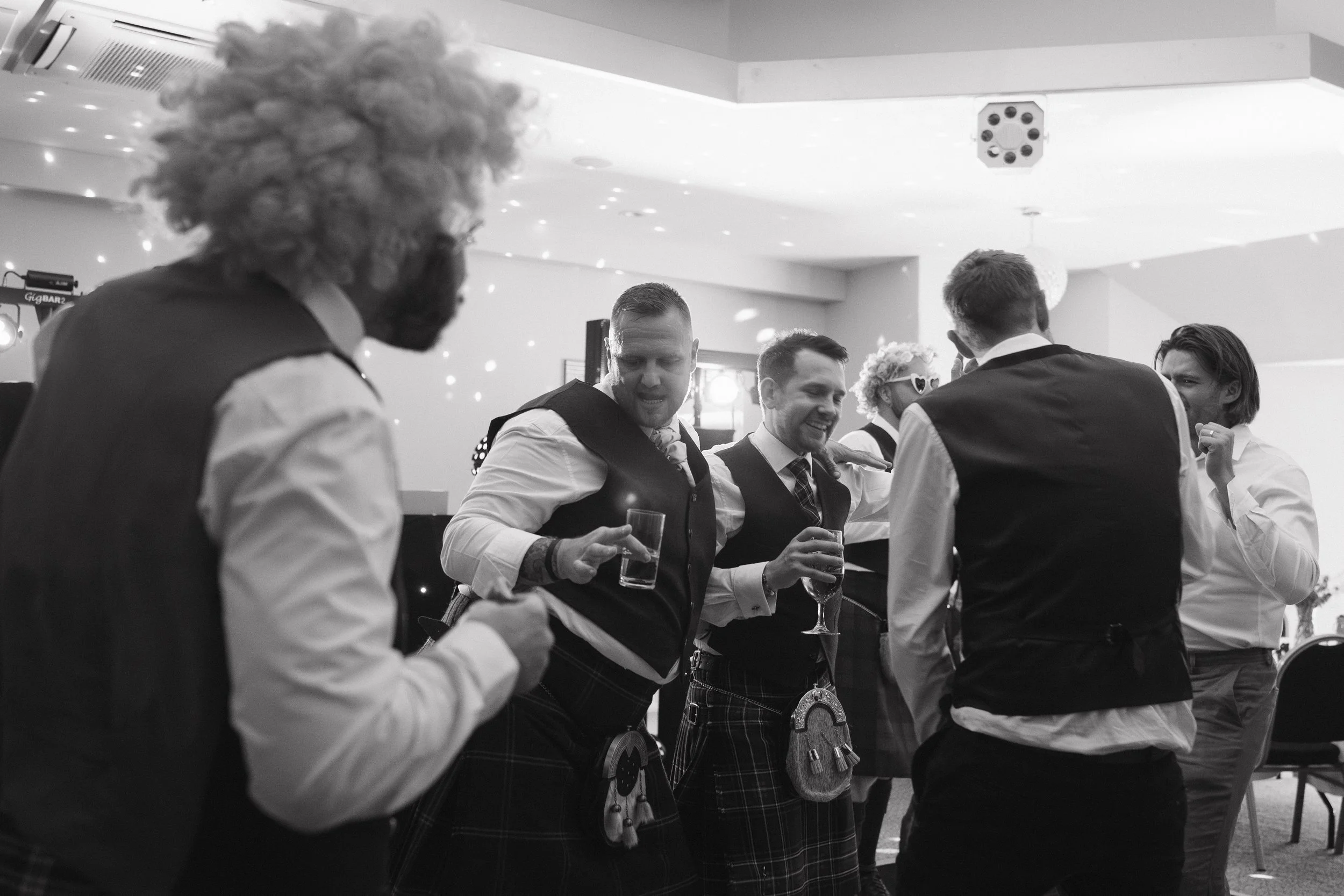 Group of men at a party, dancing and holding drinks, one wearing a clown wig and sunglasses, some wearing kilts and vests. - captured by an Edinburgh wedding photographer