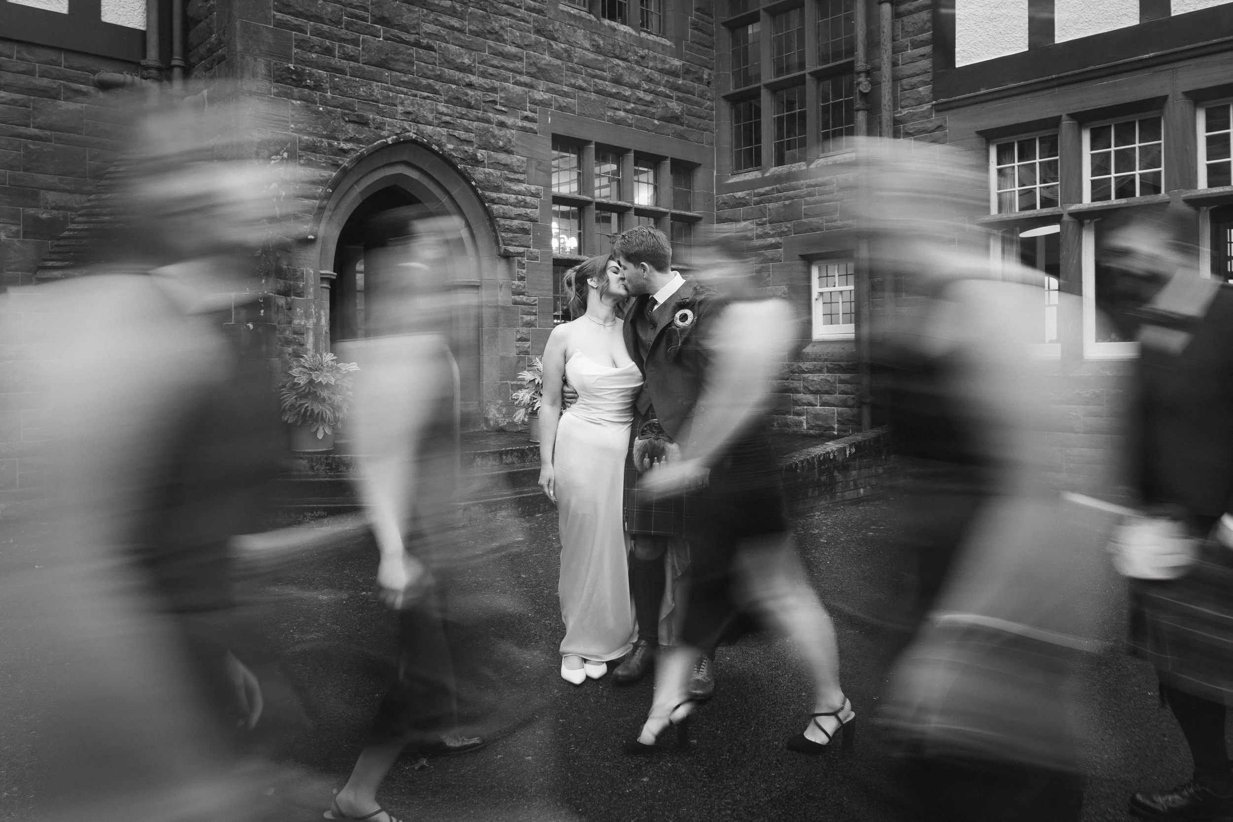 A black-and-white photo of a newlywed couple kissing in front of a historic brick building, while a group of blurred pedestrians walk by. - captured by an Edinburgh wedding photographer