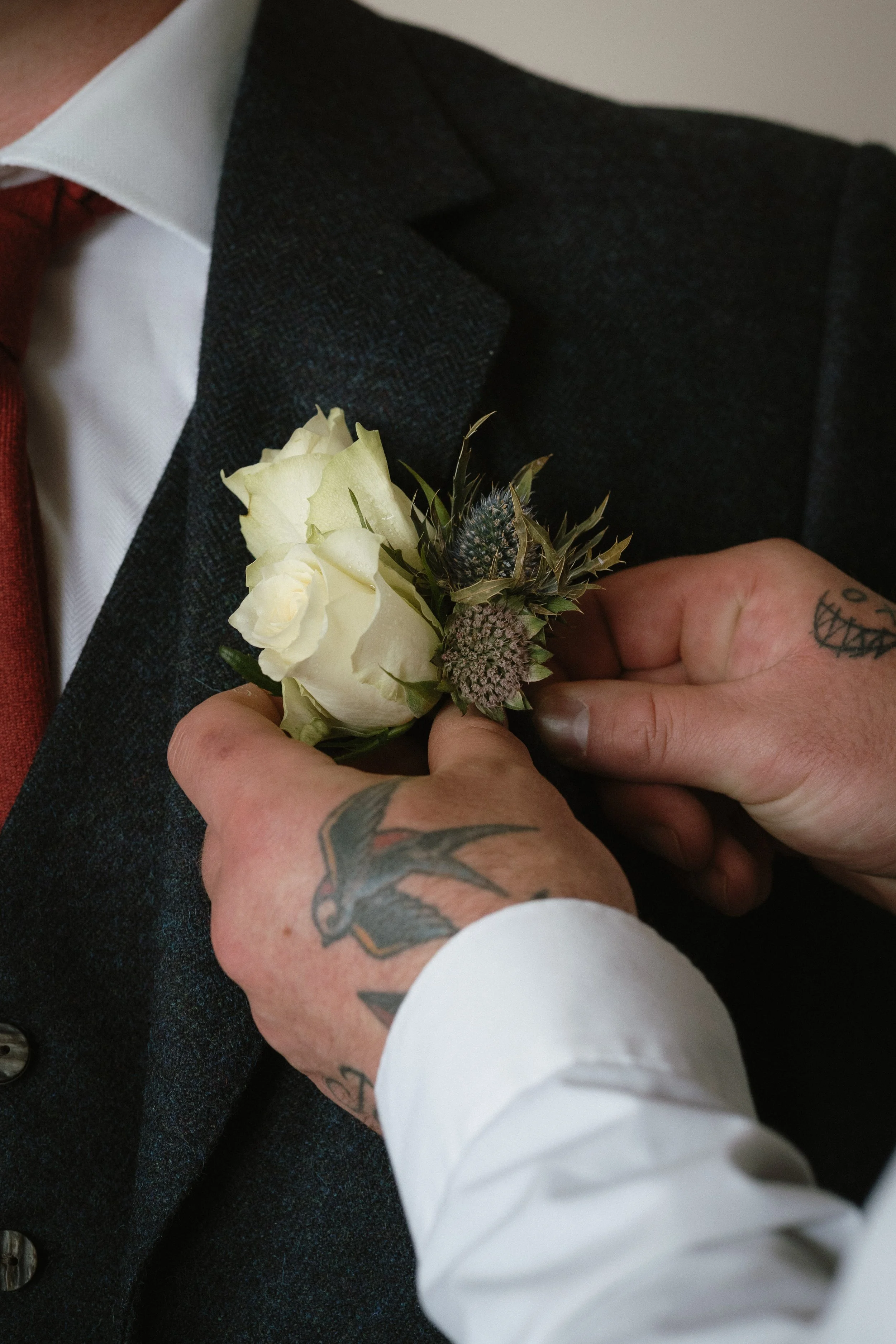 A person with tattoos on their hand has a boutonniere with white roses and greenery pinned to a dark suit jacket. - captured by an Edinburgh wedding photographer