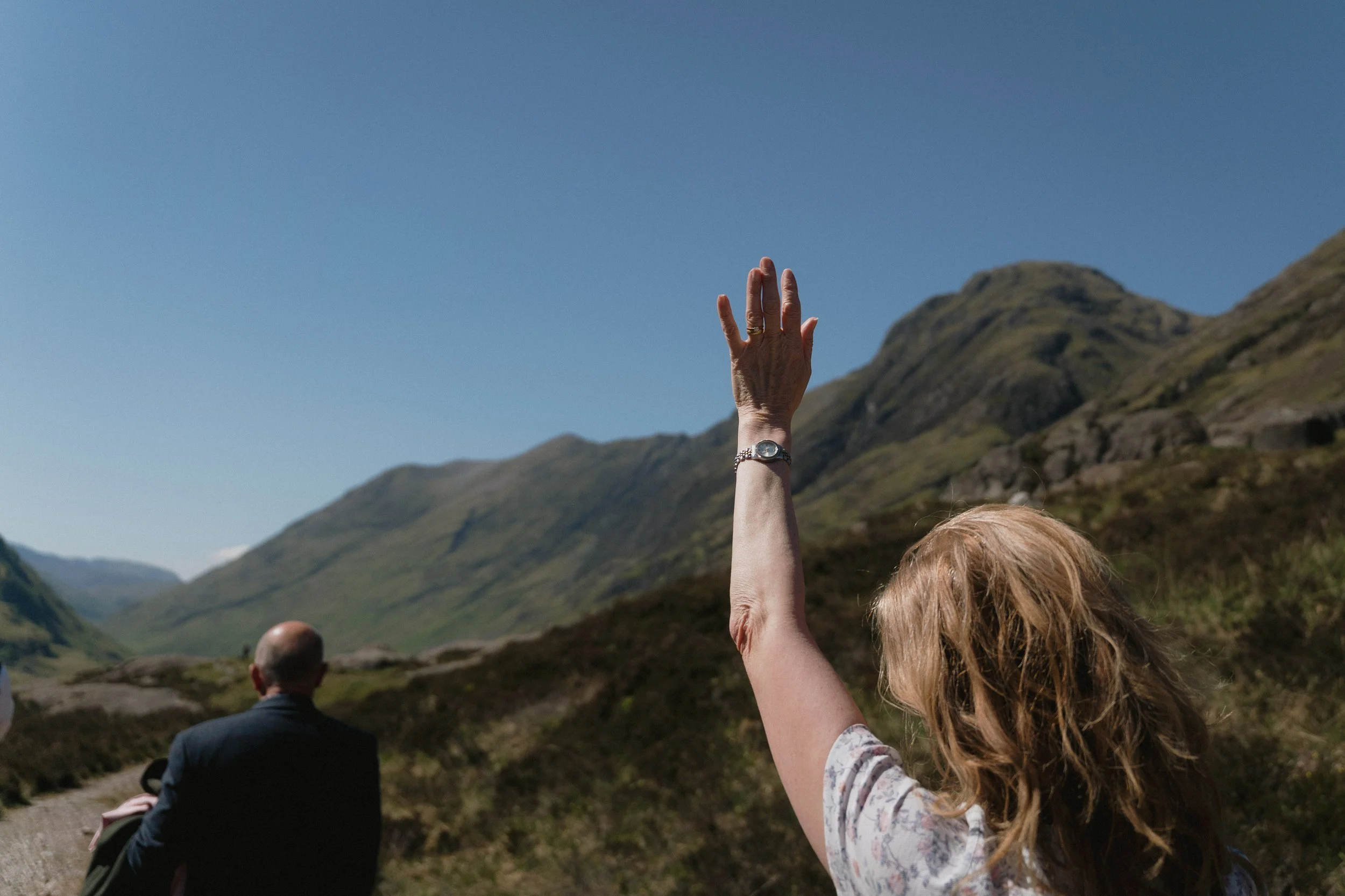 A woman with blonde hair wearing a floral top raising her hand in a mountainous landscape with clear blue sky, while a man in a dark suit sits on the ground with mountains in the background. - captured by an Edinburgh wedding photographer