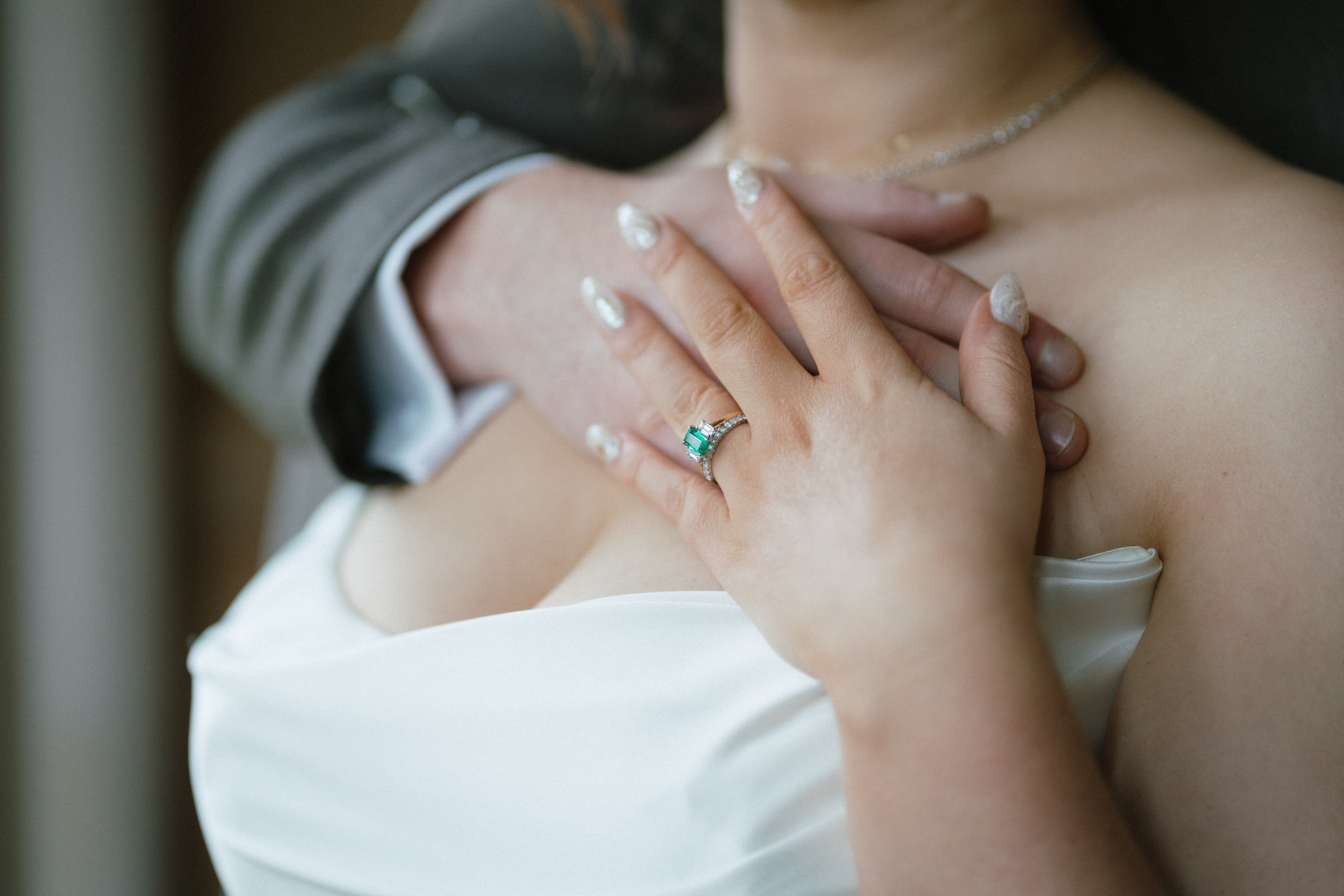 A woman with a diamond ring with a green stone on her ring finger, holding her chest with her left hand, wearing a white dress and a pearl necklace, accompanied by a man in a grey suit, whose arm is around her.