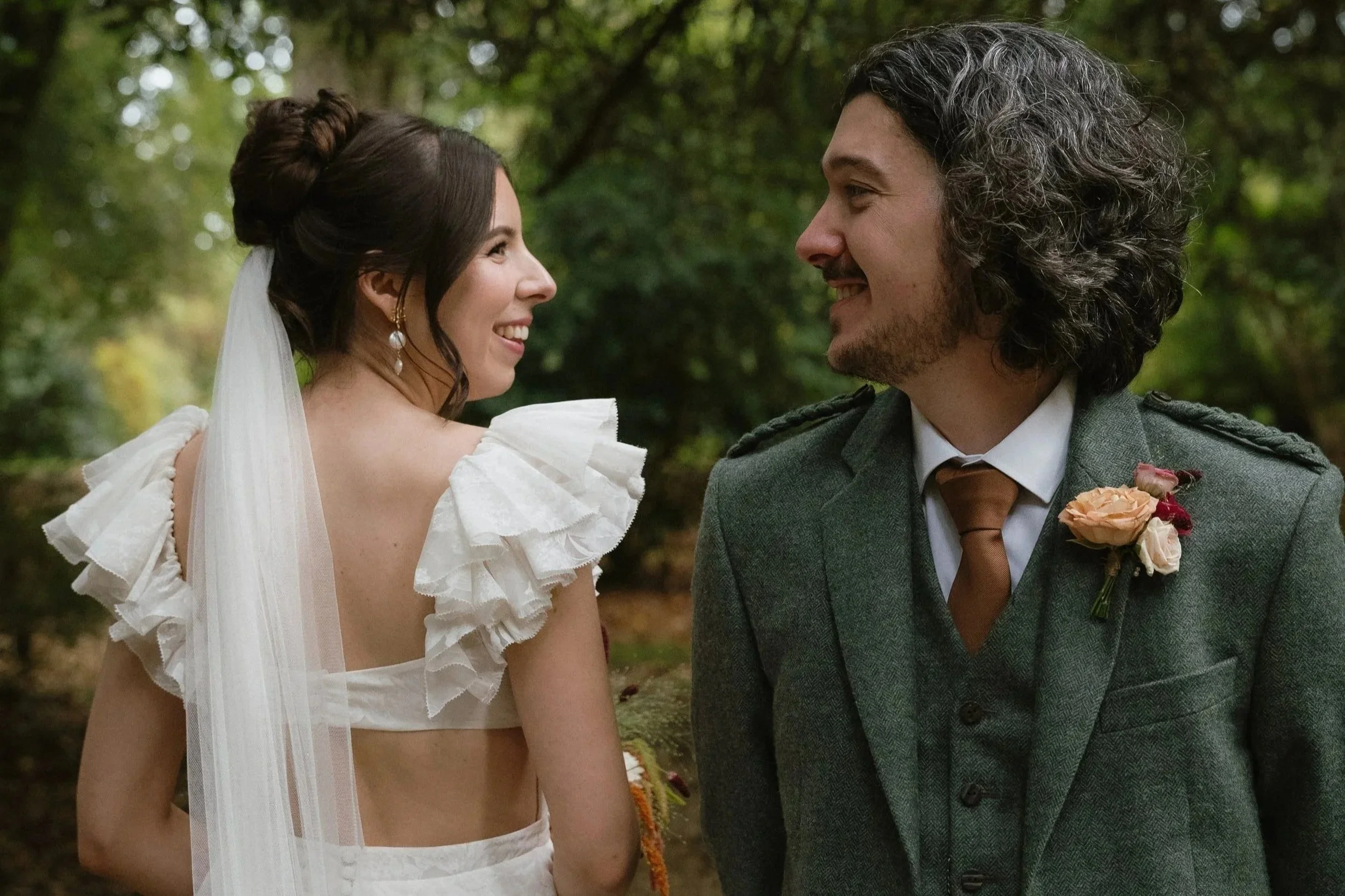 A bride and groom smiling at each other outdoors in a forested area.