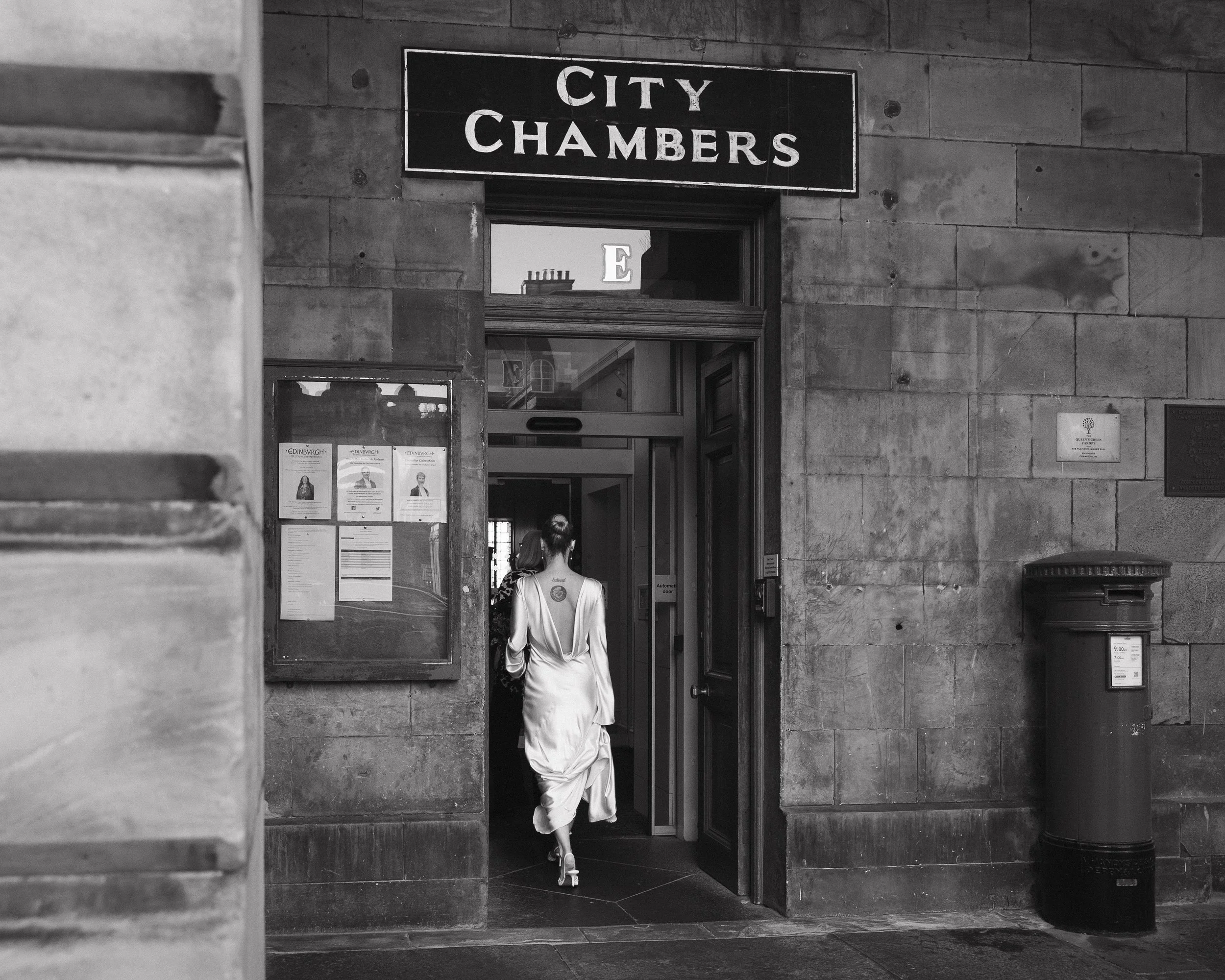 A woman in a satin dress walking into a building with a sign that reads 'City Chambers' above the entrance, with a bulletin board and a fire hydrant outside. - captured by an Edinburgh wedding photographer