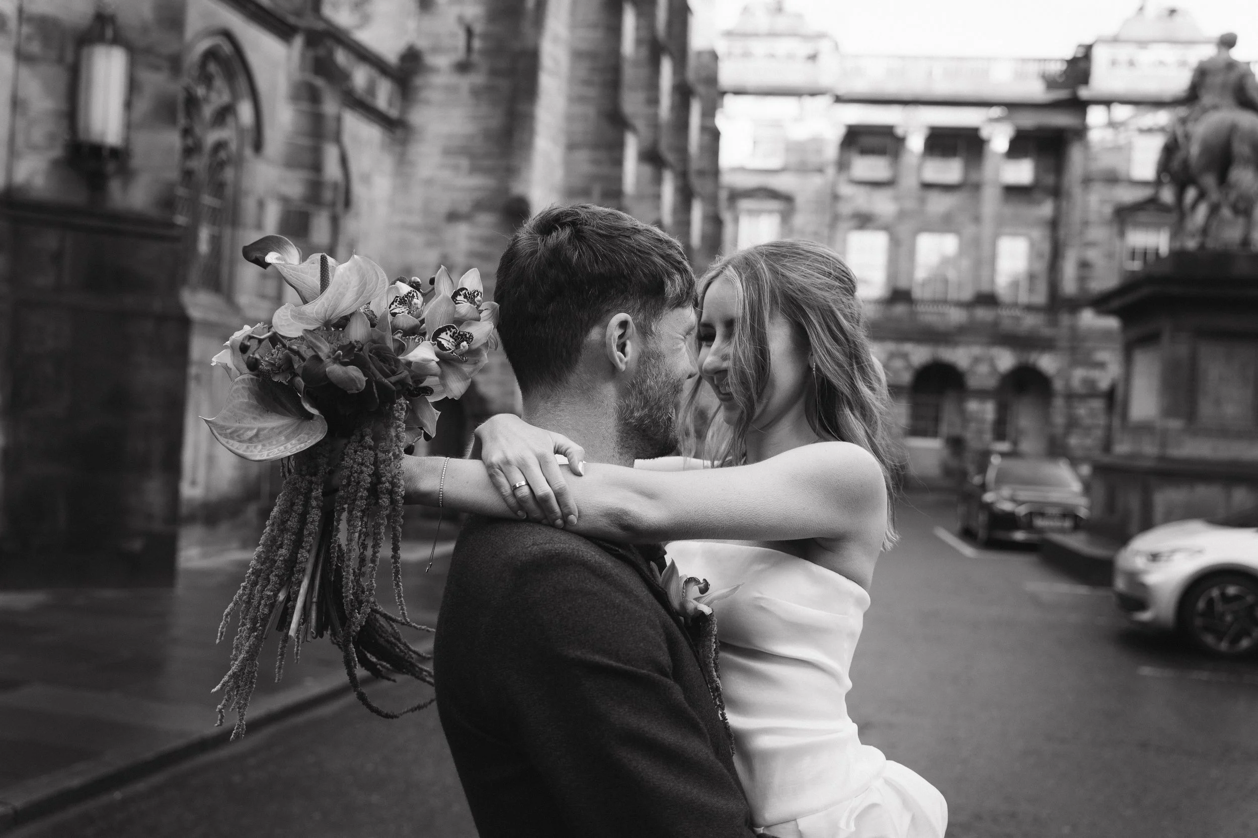 A black and white photo of a couple embracing on a city street, with historic buildings and parked cars in the background. Shot by an Edinburgh Wedding Photographer