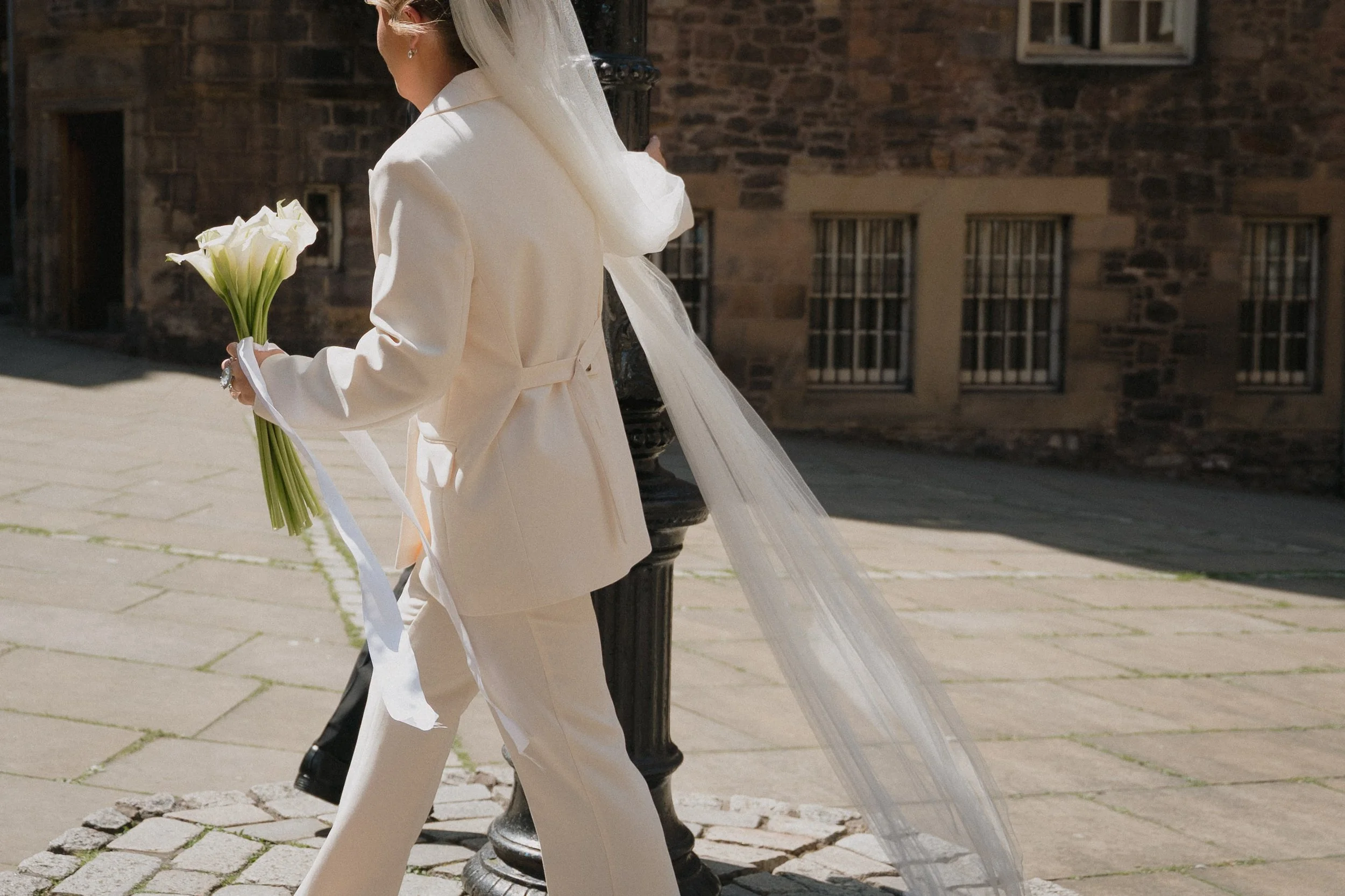 Person dressed in light-colored suit holding a bouquet of white calla lilies, standing outdoors on a stone-paved street near a brick building. - captured by an Edinburgh wedding photographer
