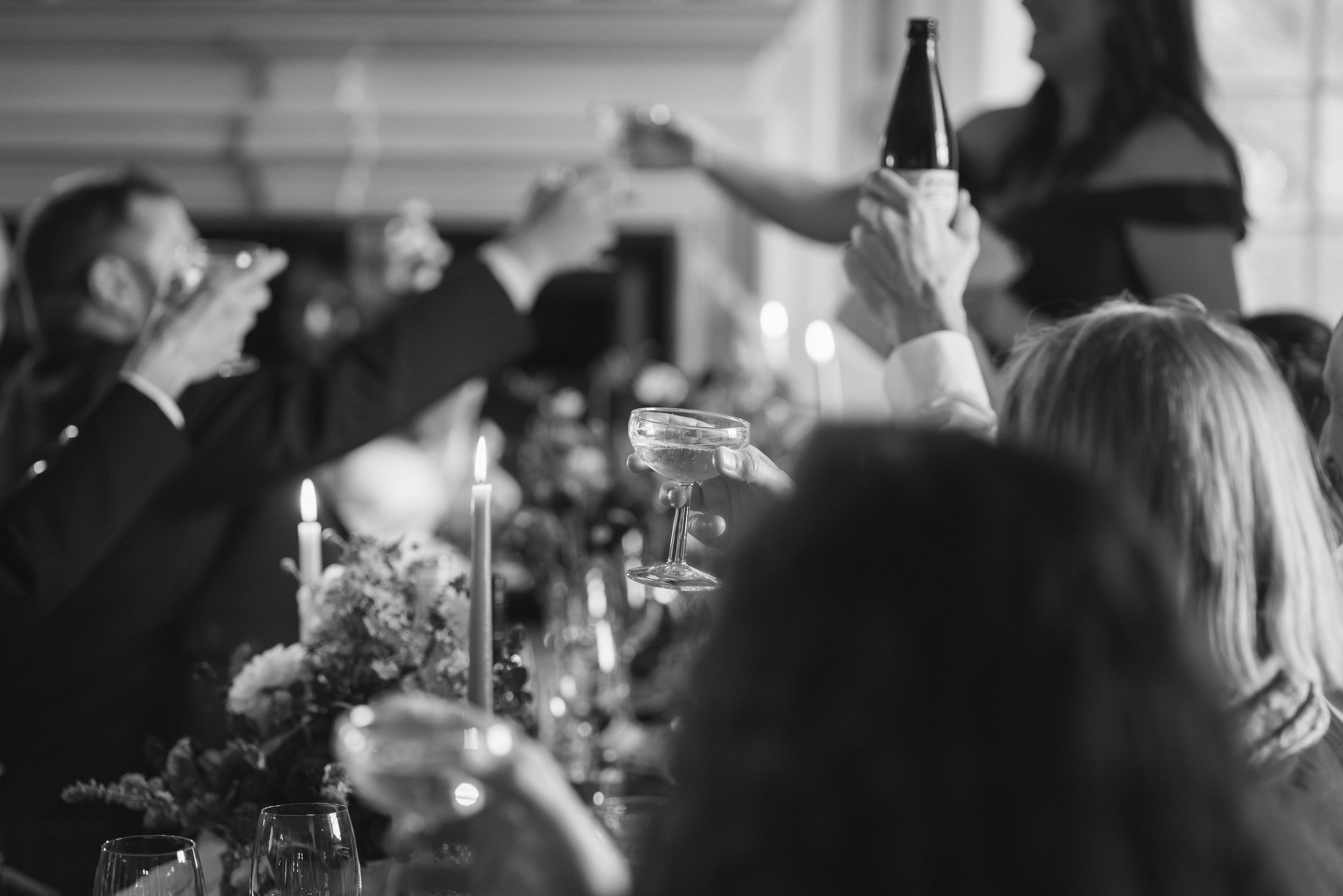 People toasting with drinks at a celebration, with a woman standing and handing out a bottle, in a festive setting. - captured by an Edinburgh wedding photographer