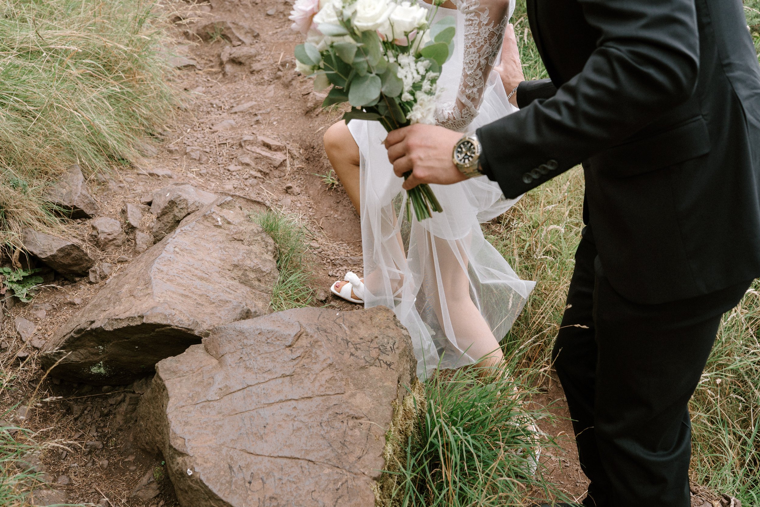 A person in a black suit holding a bouquet of flowers kneeling on a rocky dirt path next to a woman in a white lace dress, outdoors with tall grass. - captured by an Edinburgh wedding photographer
