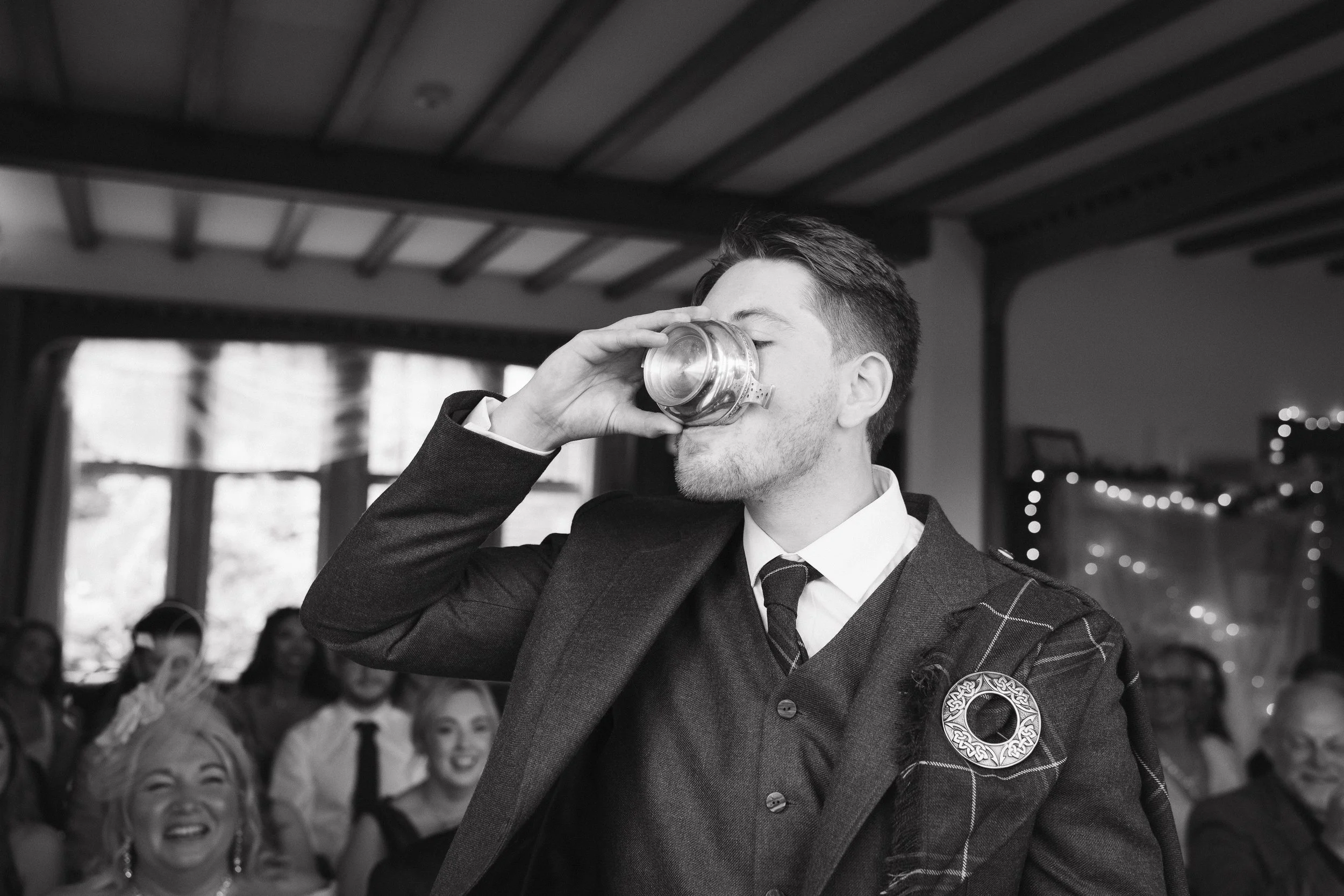 A man in formal attire, with a tartan badge on his jacket, is drinking from a glass at a social gathering. People are smiling and looking on, with a decorated background. - captured by an Edinburgh wedding photographer