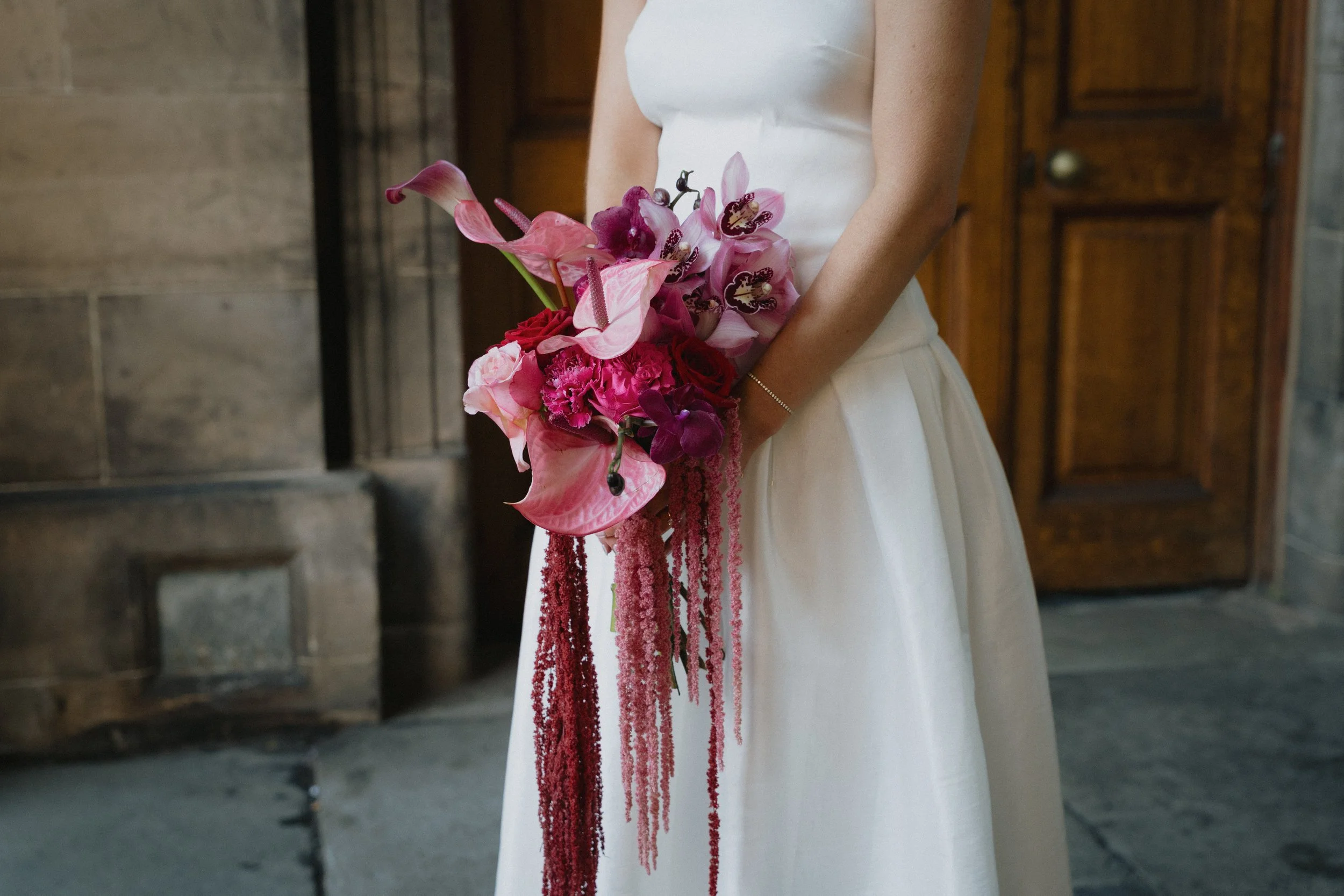 A bride in a white dress holding a bouquet of pink and purple flowers inside a stone building with a wooden door in the background. - captured by an Edinburgh wedding photographer
