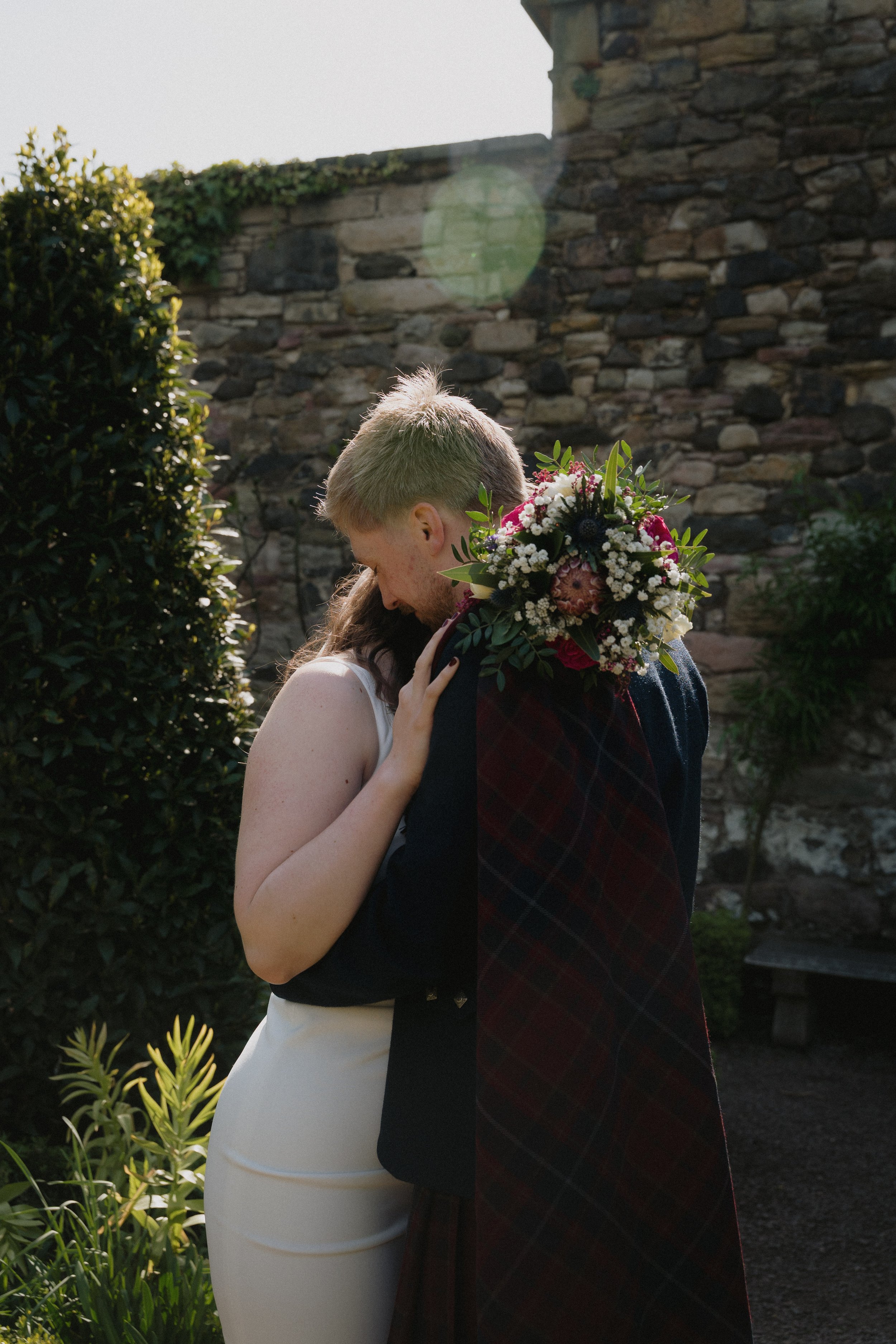 A couple embracing outdoors, with the woman holding a bouquet of colorful flowers, near a stone wall and greenery. - captured by an Edinburgh wedding photographer