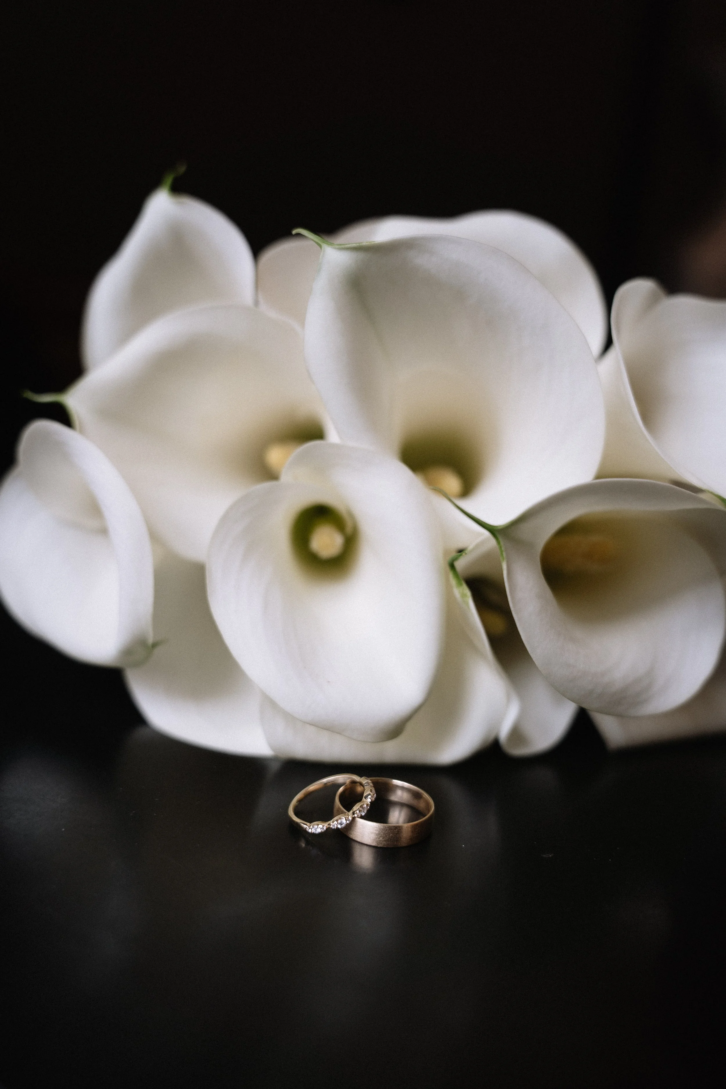 A bouquet of white calla lilies with two wedding rings placed in front on a black surface. - captured by an Edinburgh wedding photographer
