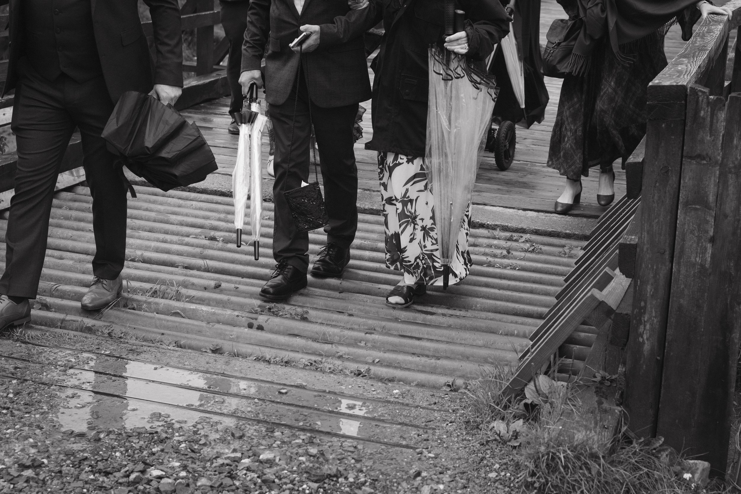 People walking on a wooden boardwalk carrying umbrellas and bags, dressed in formal and casual attire. - captured by an Edinburgh wedding photographer