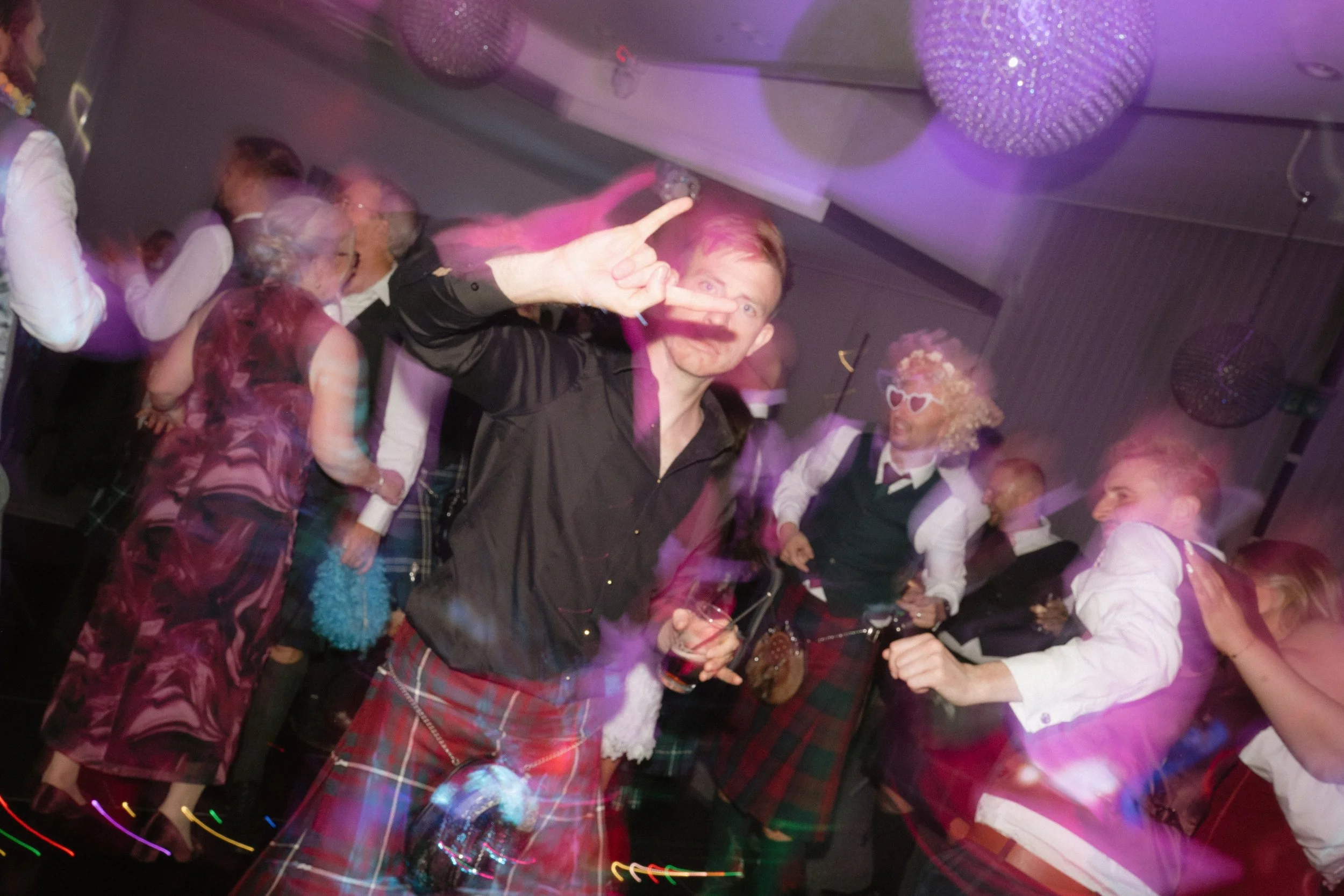 People dancing and having fun at a party, with some wearing costumes and accessories like curly wigs and sunglasses, in a dimly lit room with purple lighting. - captured by an Edinburgh wedding photographer