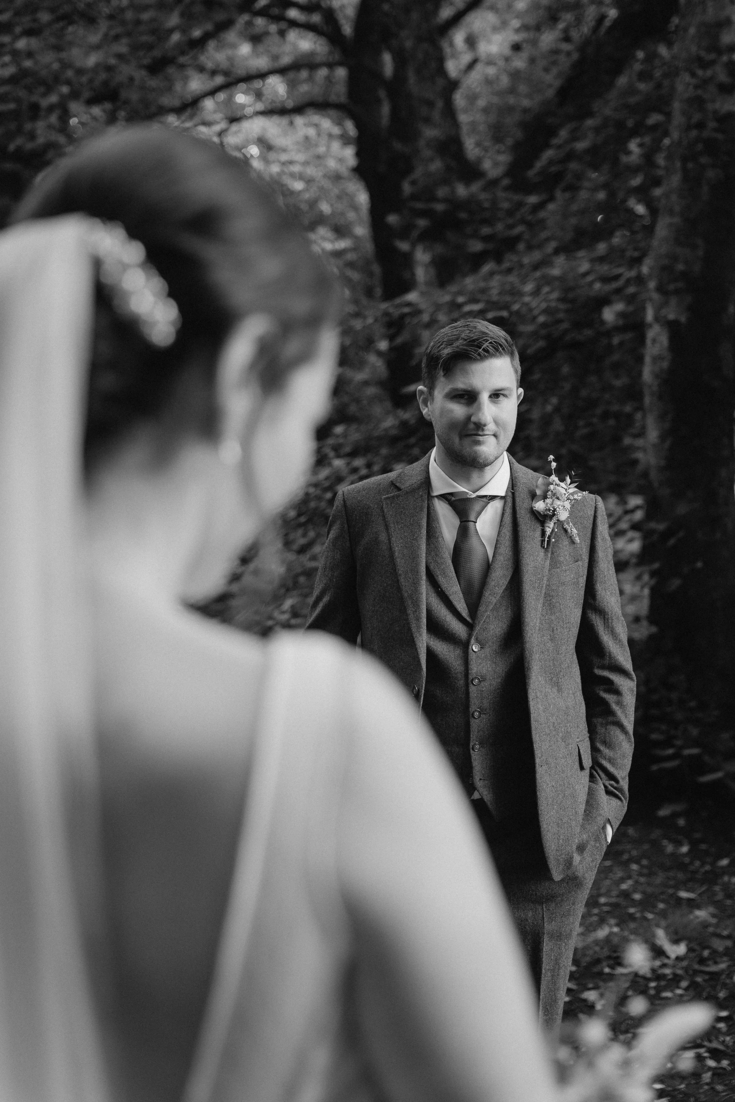 A black and white photo of a groom and bride outdoors. The groom is in the background, wearing a suit with a boutonniere. The bride is in the foreground, slightly out of focus, with her hair in an updo. - captured by an Edinburgh wedding photographer