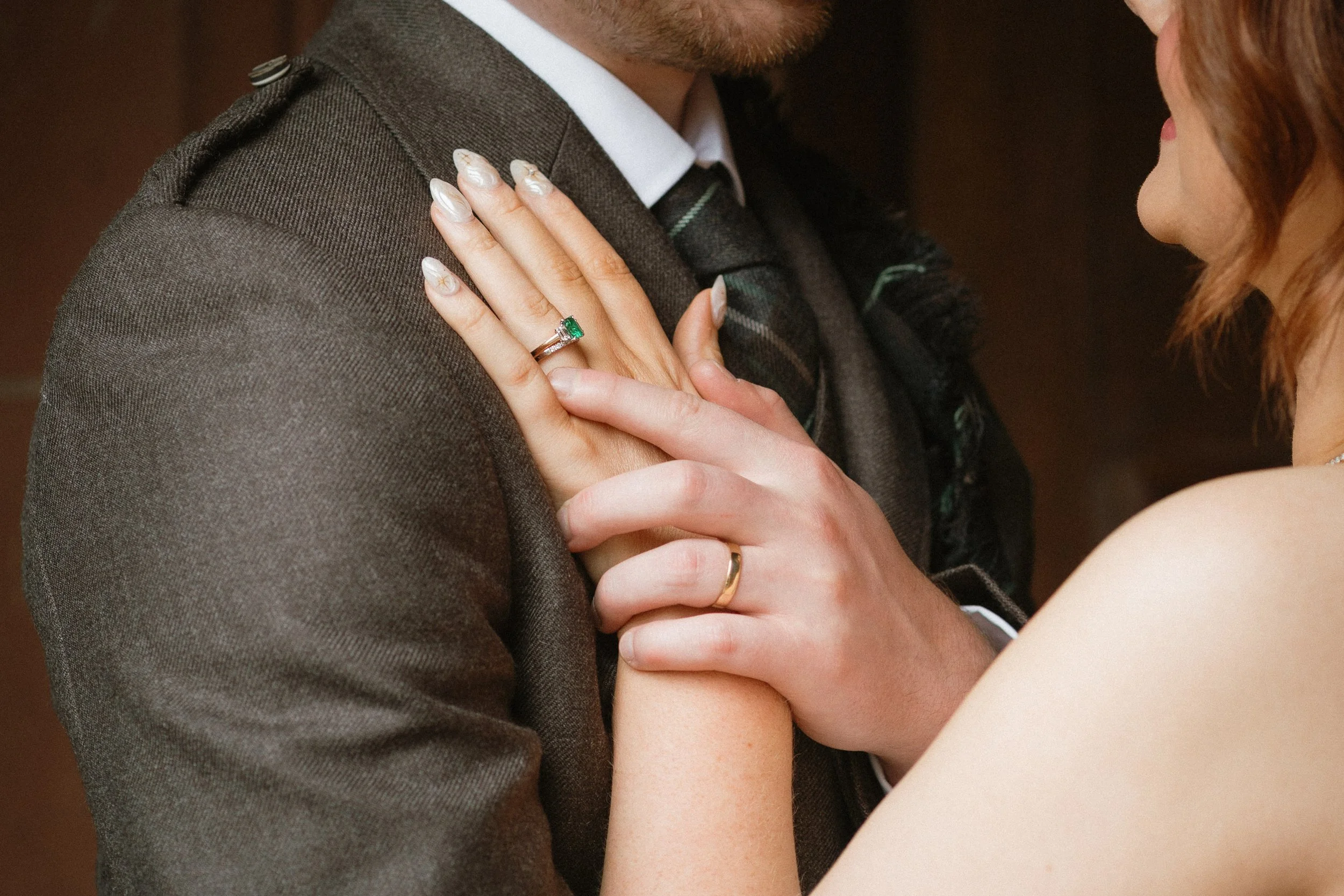 A close-up of a couple's hands and torsos, with their rings visible; the woman is wearing a ring with an emerald gemstone, and both are embracing each other. - captured by an Edinburgh wedding photographer