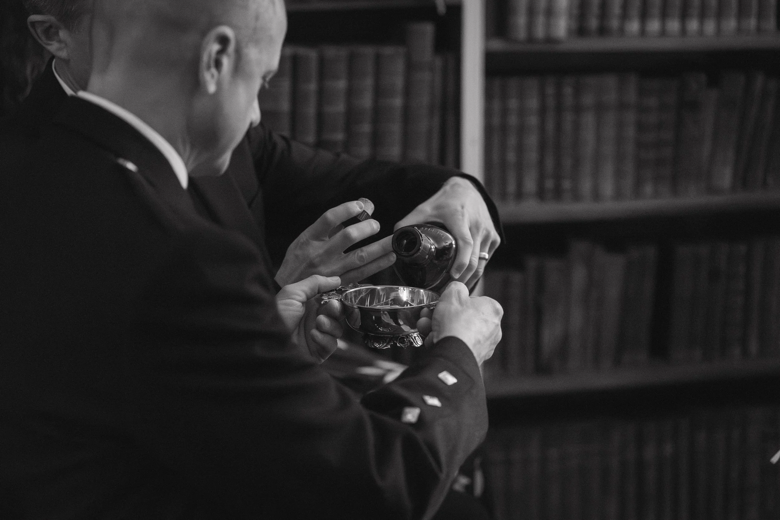 A person pouring a drink from a bottle into a small bowl, with bookshelves in the background. - captured by an Edinburgh wedding photographer