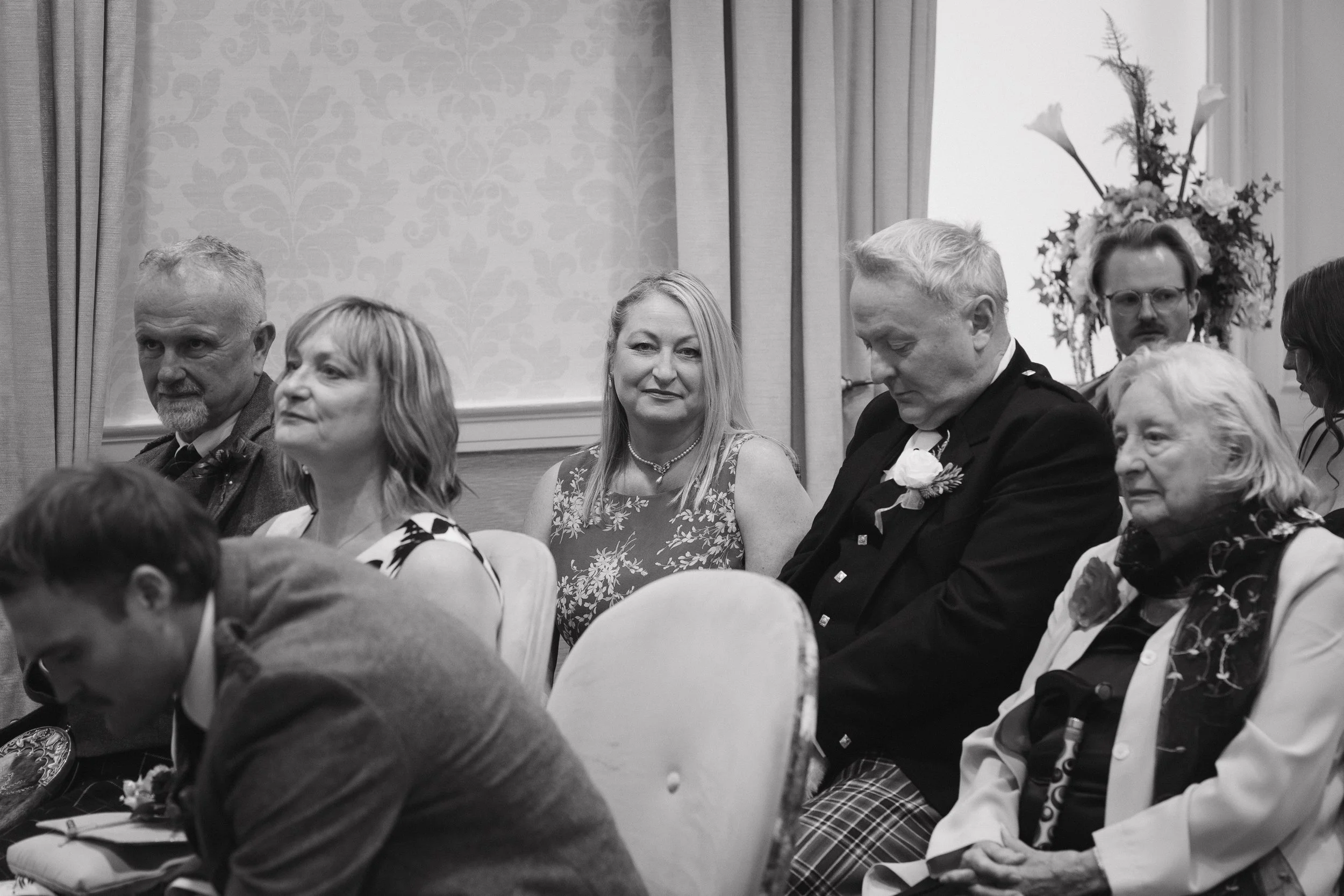 Black and white photo of a group of people sitting in a room, some wearing formal attire, at what appears to be a social or formal event. - captured by an Edinburgh wedding photographer