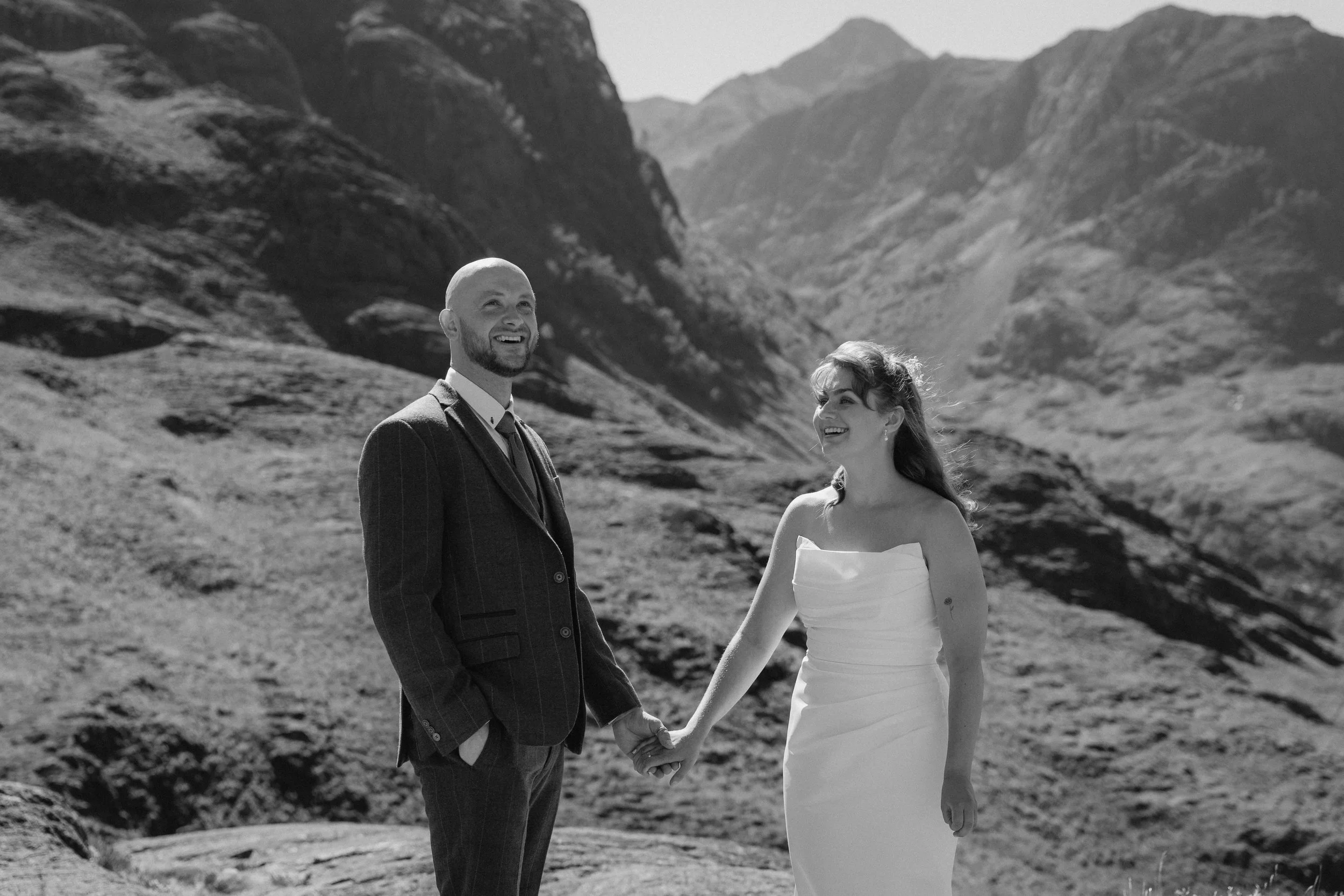 A bride and groom holding hands and smiling at each other outdoors in a mountainous landscape. - captured by an Edinburgh wedding photographer