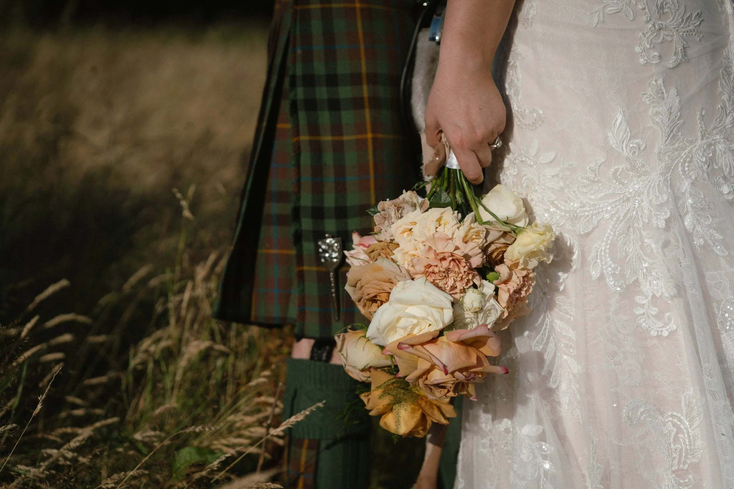 Bride holding a bouquet of flowers, wearing a white lace wedding dress, and standing next to a person dressed in traditional Scottish kilt attire. - captured by an Edinburgh wedding photographer