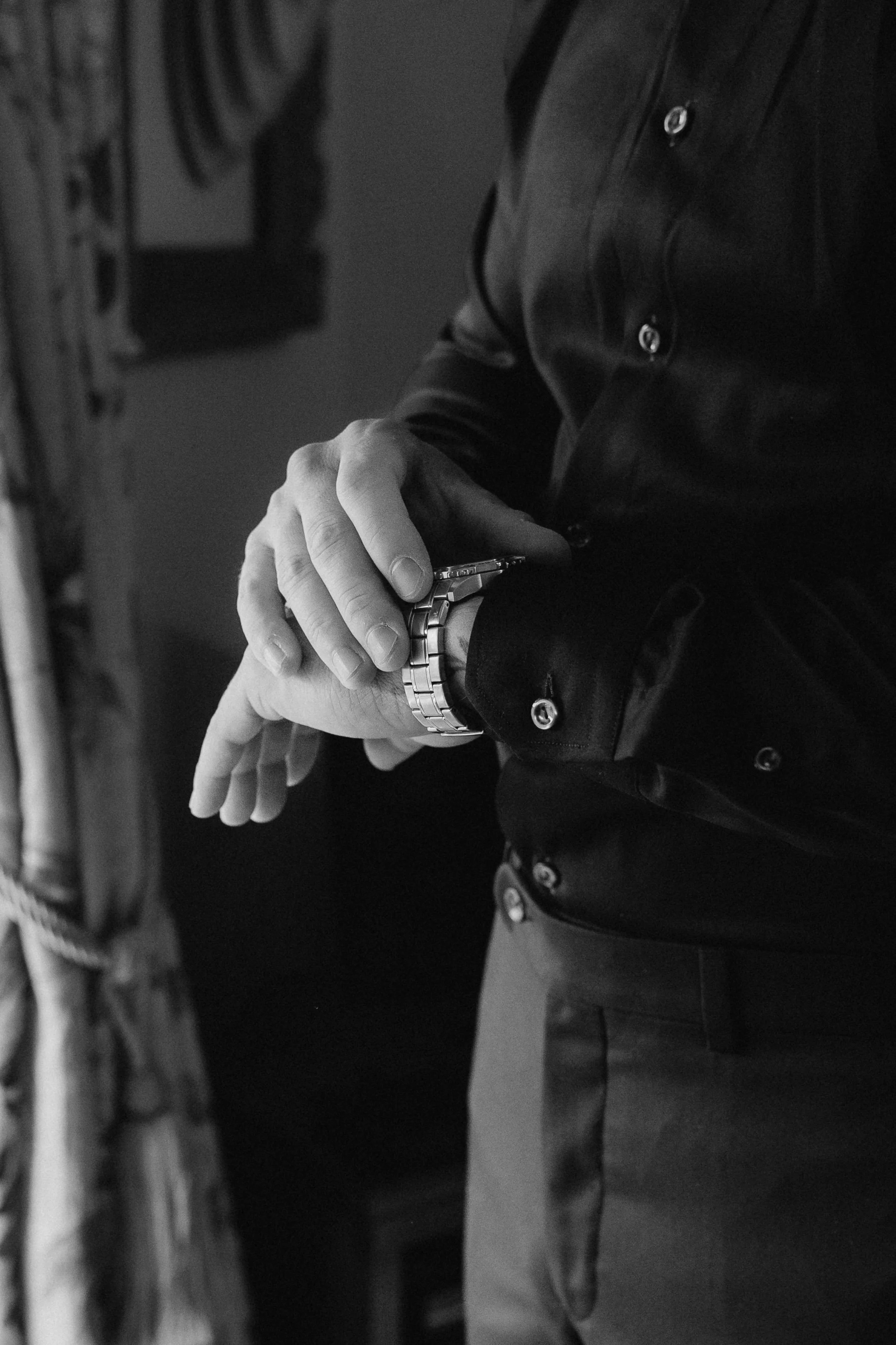 Black and white photo of a man wearing a long-sleeve dark shirt with rolled-up sleeves, adjusting his watch. - captured by an Edinburgh wedding photographer