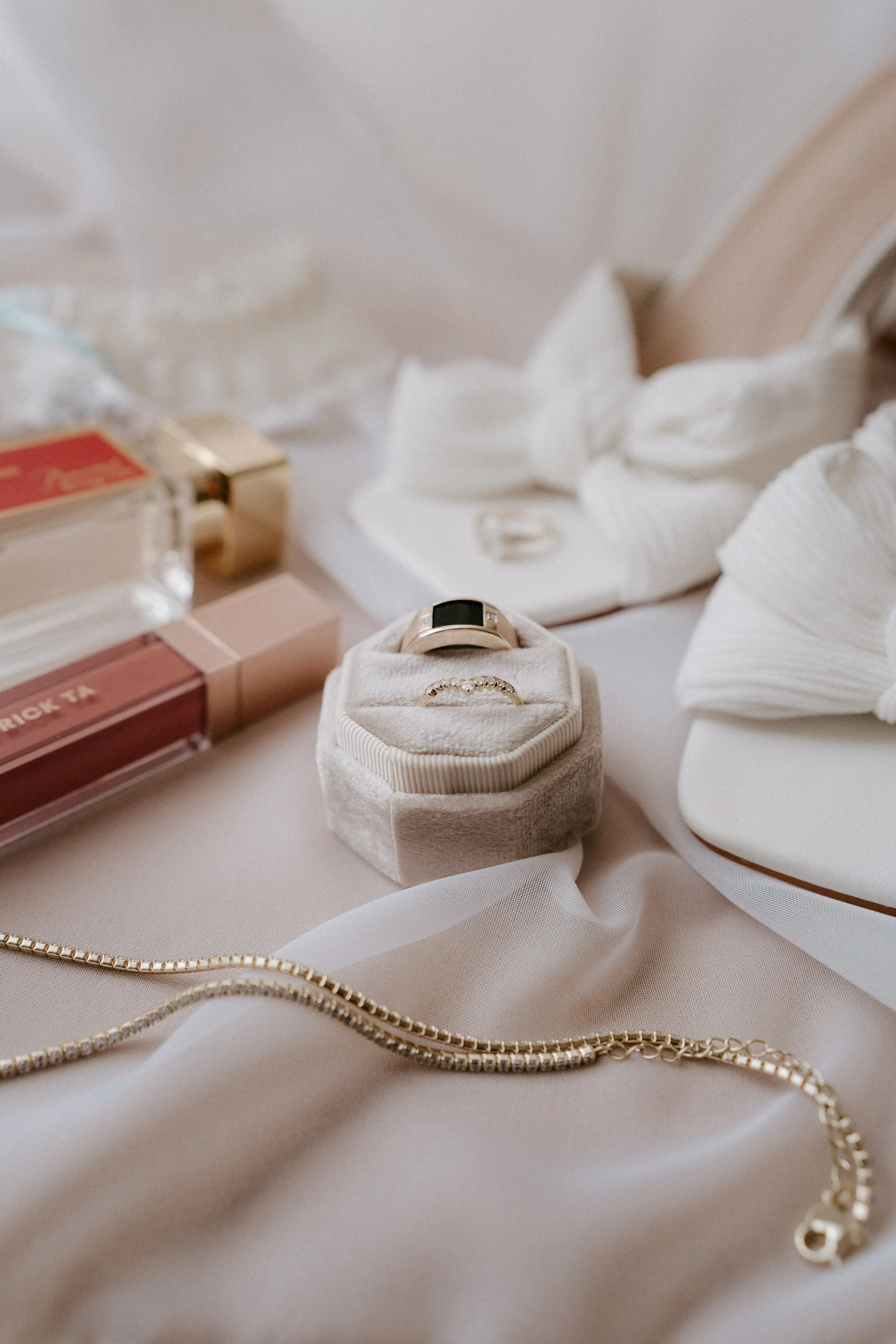 Close-up of jewelry and beauty products on a soft beige surface, including a gold necklace, a ring in a cream-colored ring box, a smartwatch, and lip gloss, with decorative white seashells and tissue paper.