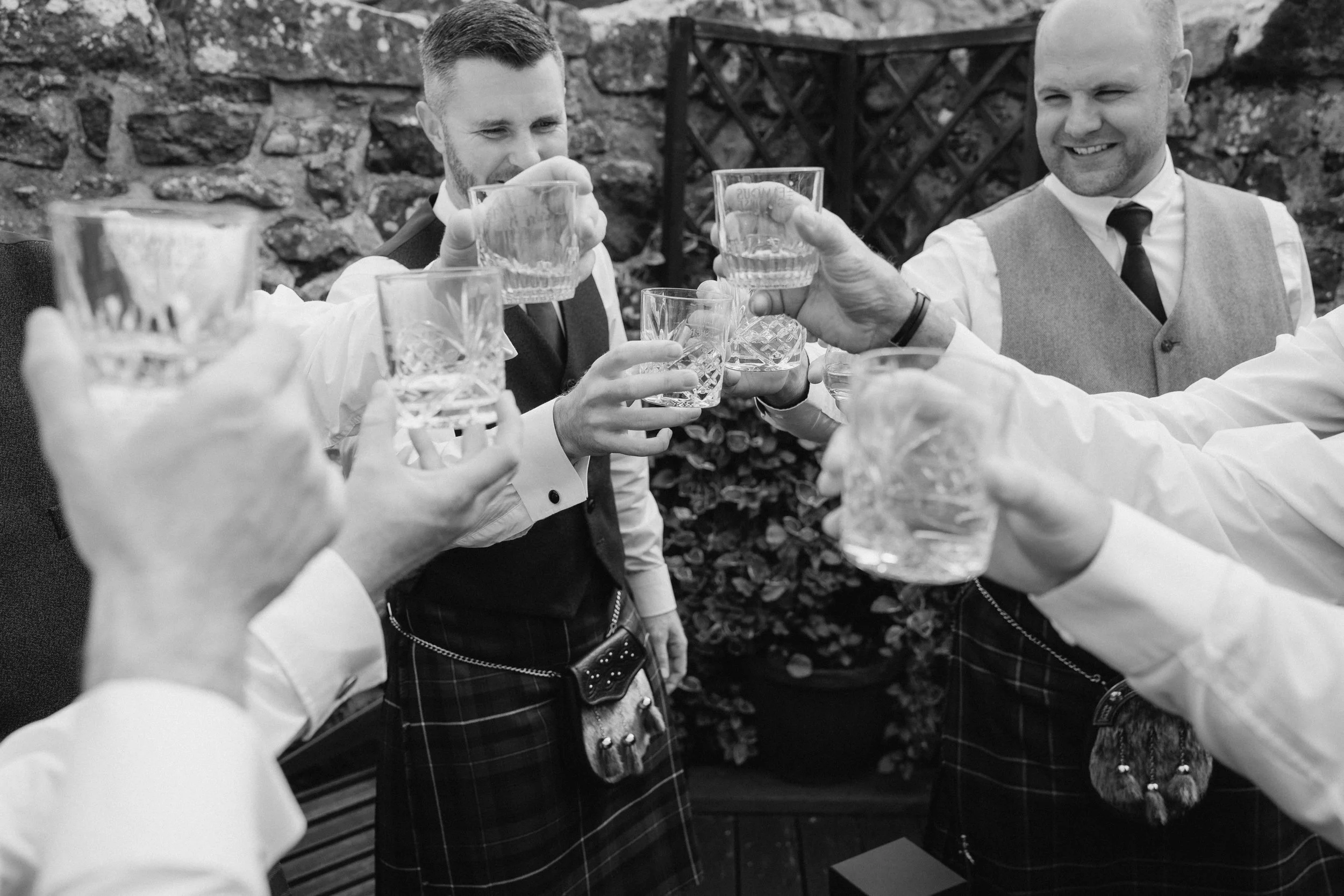 A group of men dressed in traditional Scottish attire, including kilts, raising glasses for a toast outdoors with a stone wall in the background. - captured by an Edinburgh wedding photographer