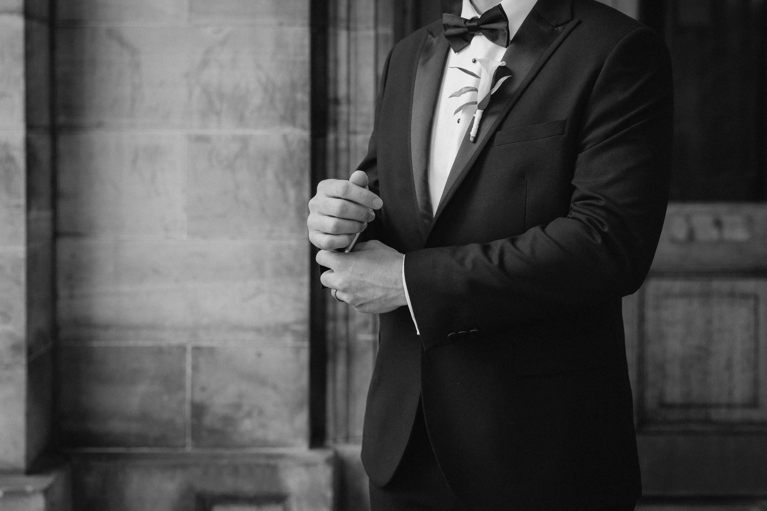 A man in a black tuxedo, white dress shirt, and black bow tie adjusting his dress cuff. - captured by an Edinburgh wedding photographer
