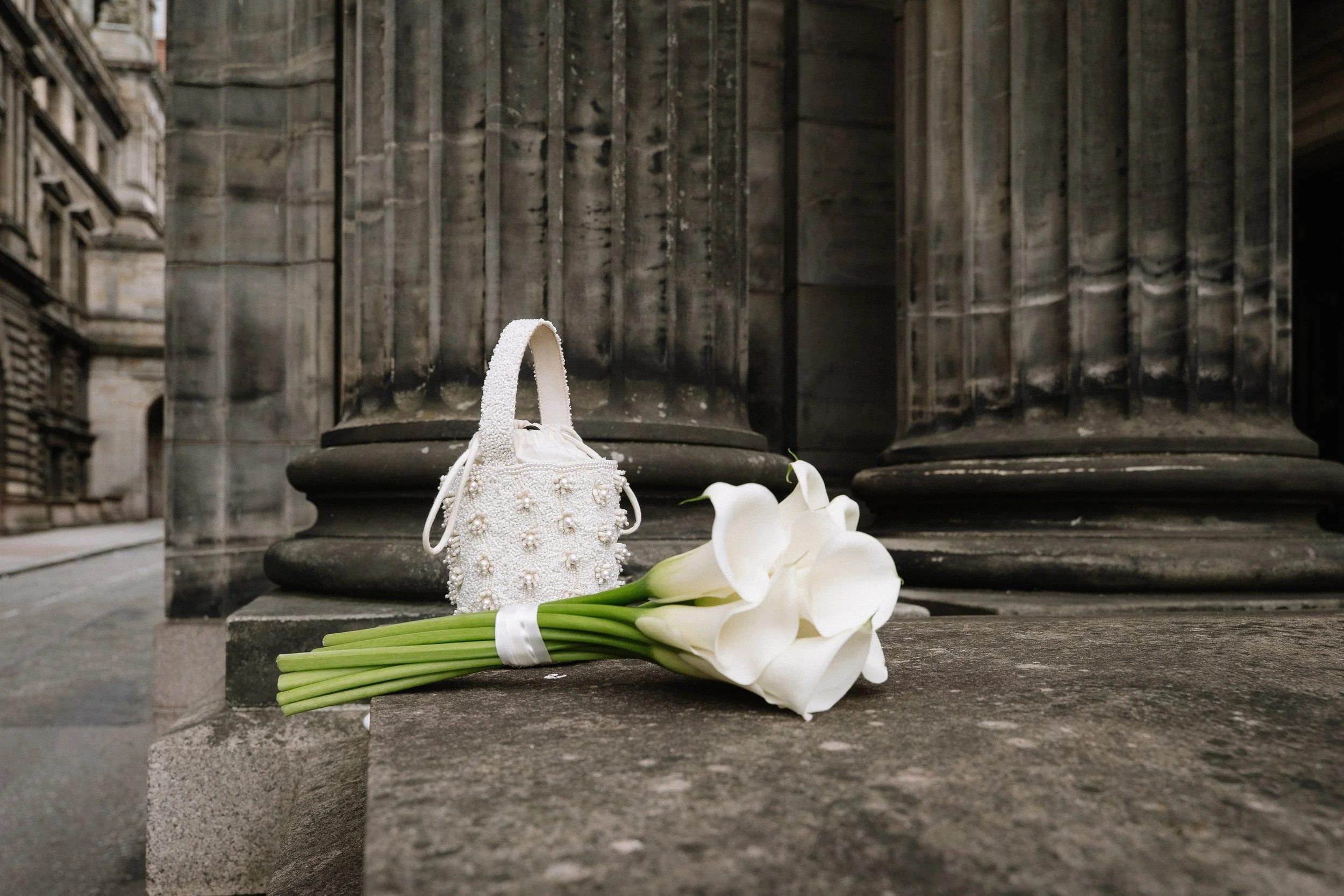 A bouquet of white calla lilies and a white handbag with pearls and flower embellishments are placed on a stone step against large stone columns. - captured by an Edinburgh wedding photographer