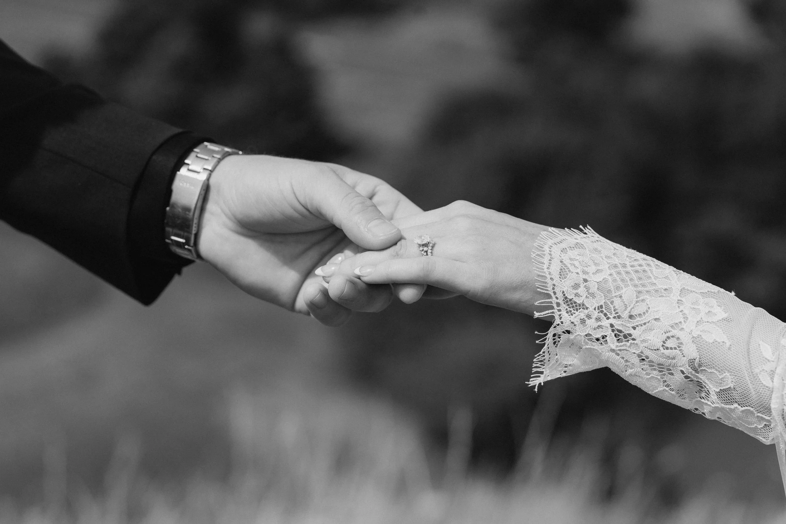 Close-up of a groom holding the bride's hand, showing her wedding ring, with her wearing lace wedding dress, in black and white.