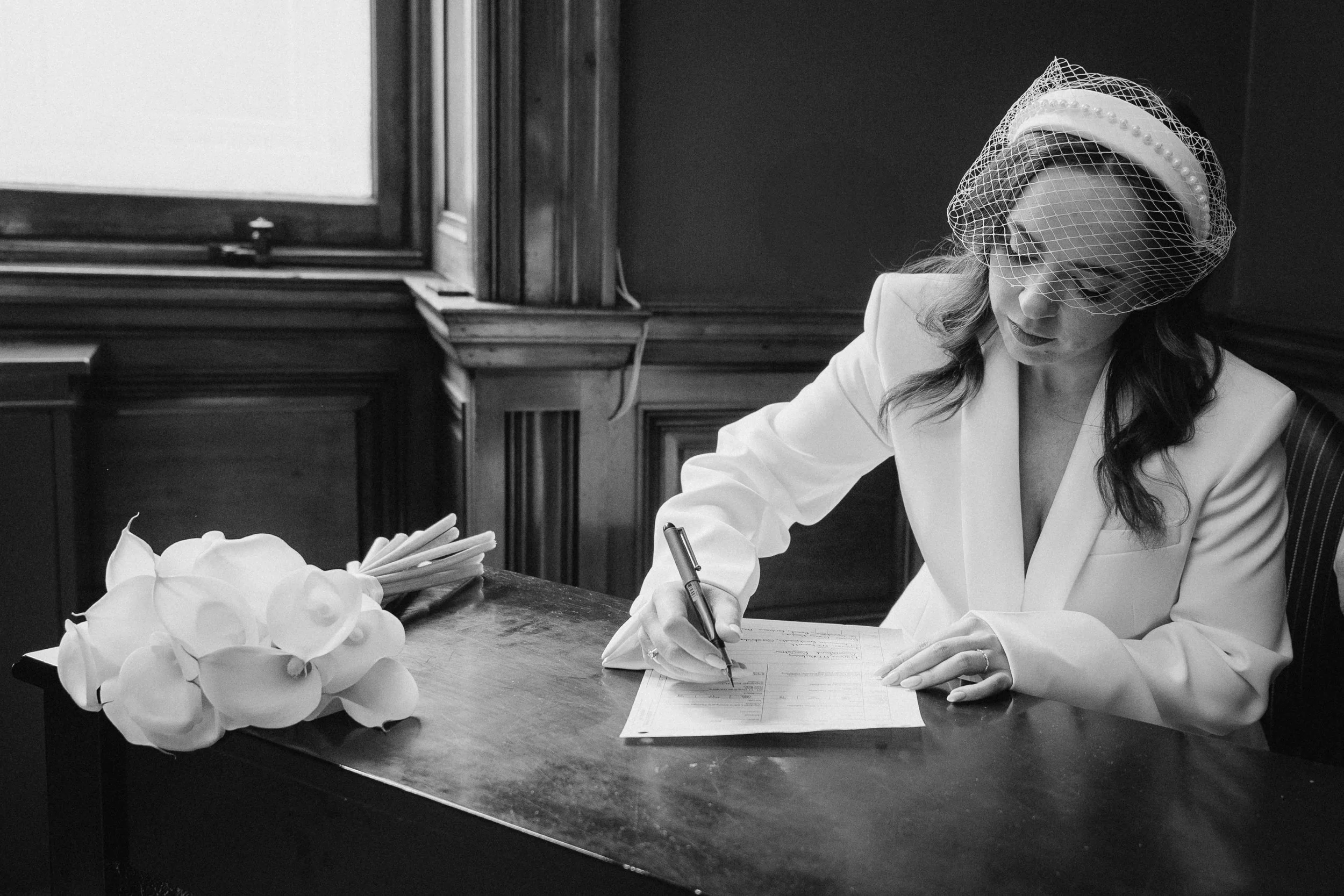 A woman in a white suit signing a document at a wooden table, wearing a birdcage veil and a headband, with a bouquet of flowers placed on the table. - captured by an Edinburgh wedding photographer