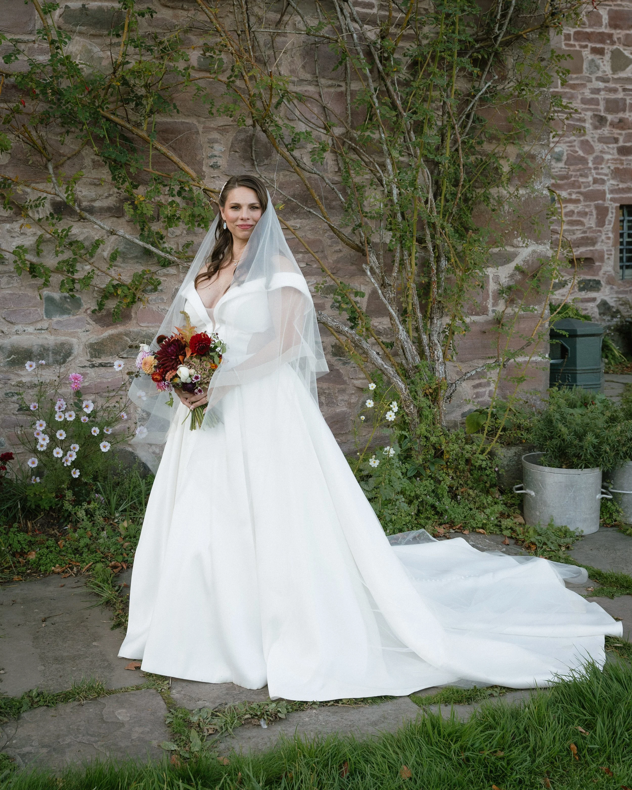 Bride in a white wedding dress holding a bouquet outdoors in front of a brick wall and greenery. - captured by an Edinburgh wedding photographer
