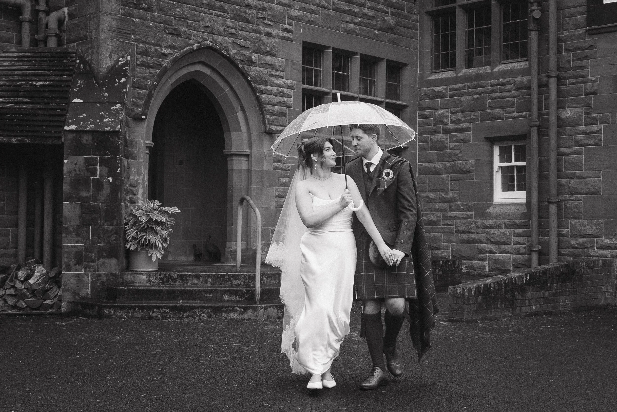 A bride and groom holding hands under an umbrella outside a stone building, gazing at each other, in black and white. - captured by an Edinburgh wedding photographer