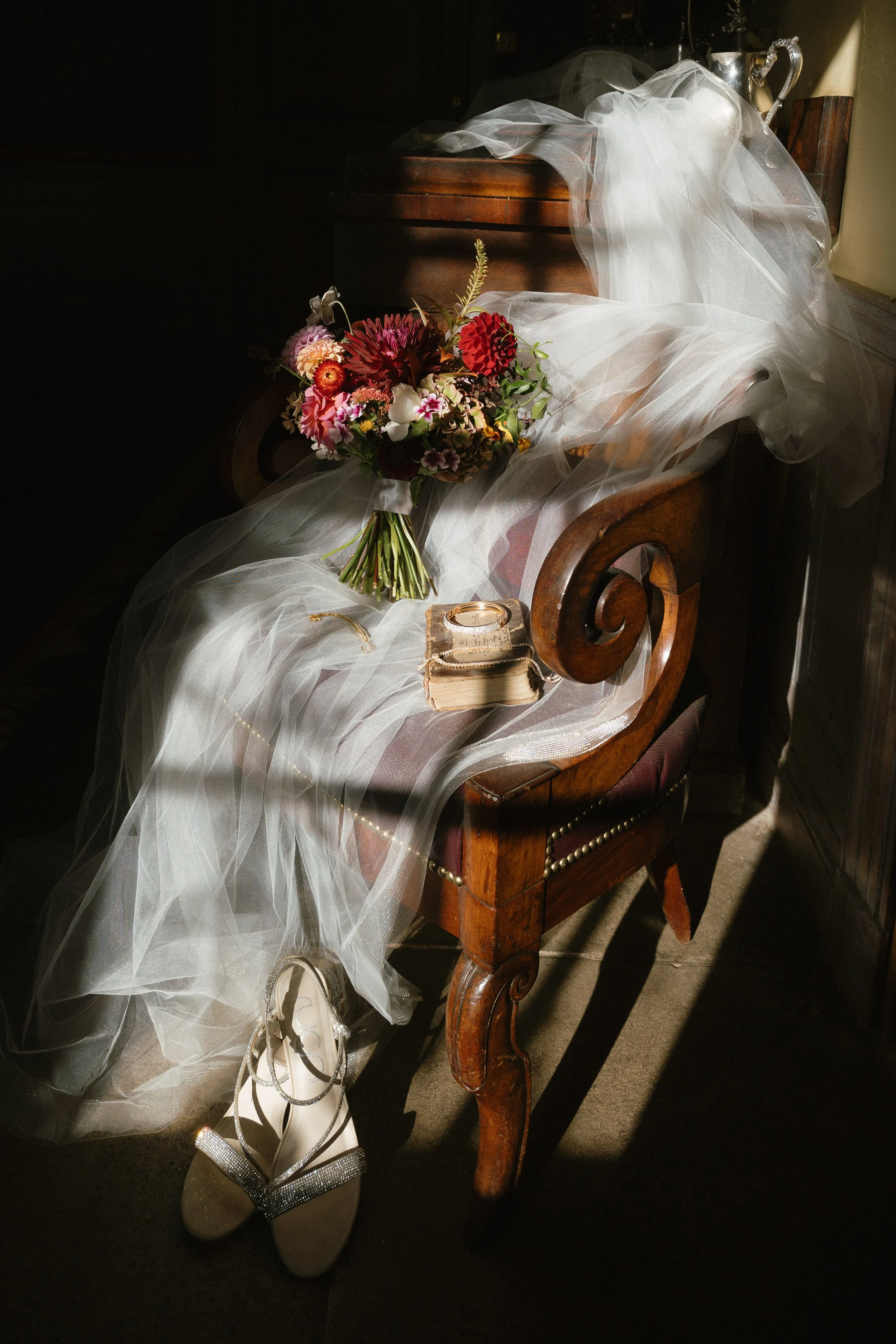 A wooden bench with a veil, a bouquet of colorful flowers, a pair of white shoes with ankle straps, and a small box with a wedding ring on top, all illuminated by soft sunlight. - captured by an Edinburgh wedding photographer