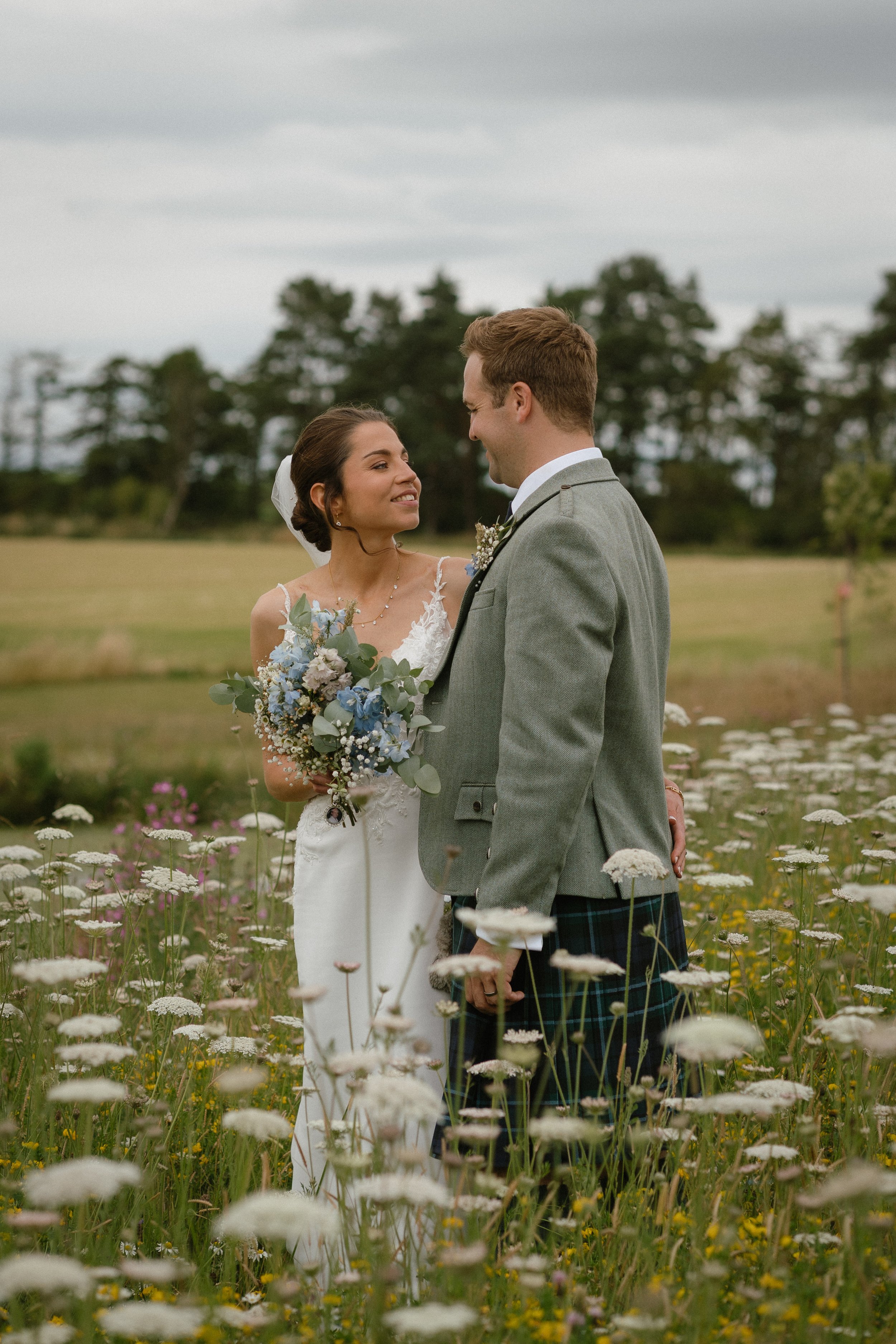 Bride and groom stand close in a field of wildflowers, gazing into each other's eyes against a cloudy sky background - captured by an Edinburgh wedding photographer