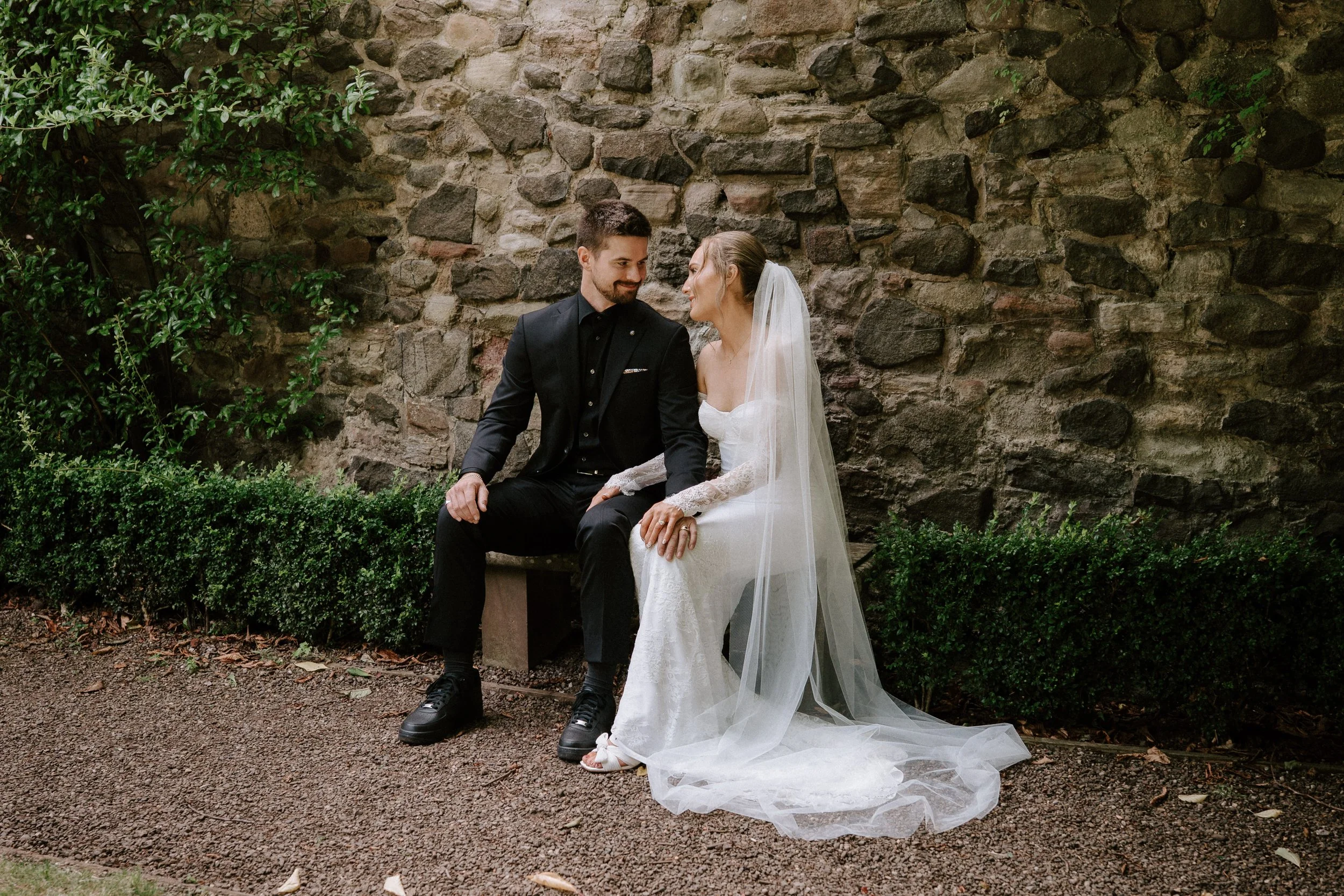 A bride and groom sitting on a bench, looking at each other, in front of a stone wall. - captured by an Edinburgh wedding photographer